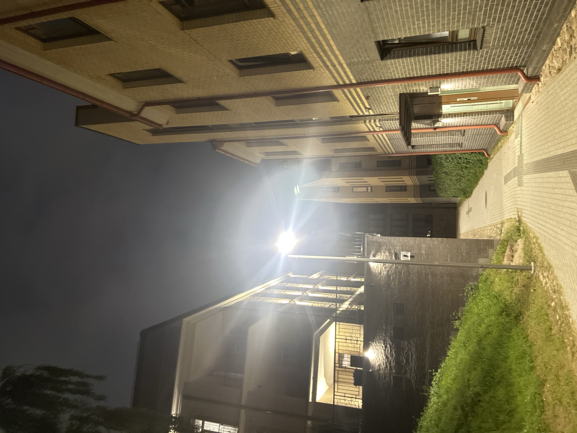 The image depicts a nighttime scene in a city with a dark sky overhead. In the foreground, a paved pathway with textured lines runs between two buildings. To the left, a building with a dark facade and balconies with railings is illuminated from within, casting a warm glow. A grassy slope covered in green vegetation rises from the pathway towards this building. To the right, a building constructed of light brown and grey bricks features several windows and a small overhang above a doorway. Red pipes run vertically along the brick facade. In the middle ground, a street lamp with a bright light is positioned along the pathway, illuminating the scene. Another building, taller and with a similar brick construction, is visible further down the pathway. The overall impression is that of an urban alleyway or courtyard at night. The location is identified as Vilnius, Lithuania.