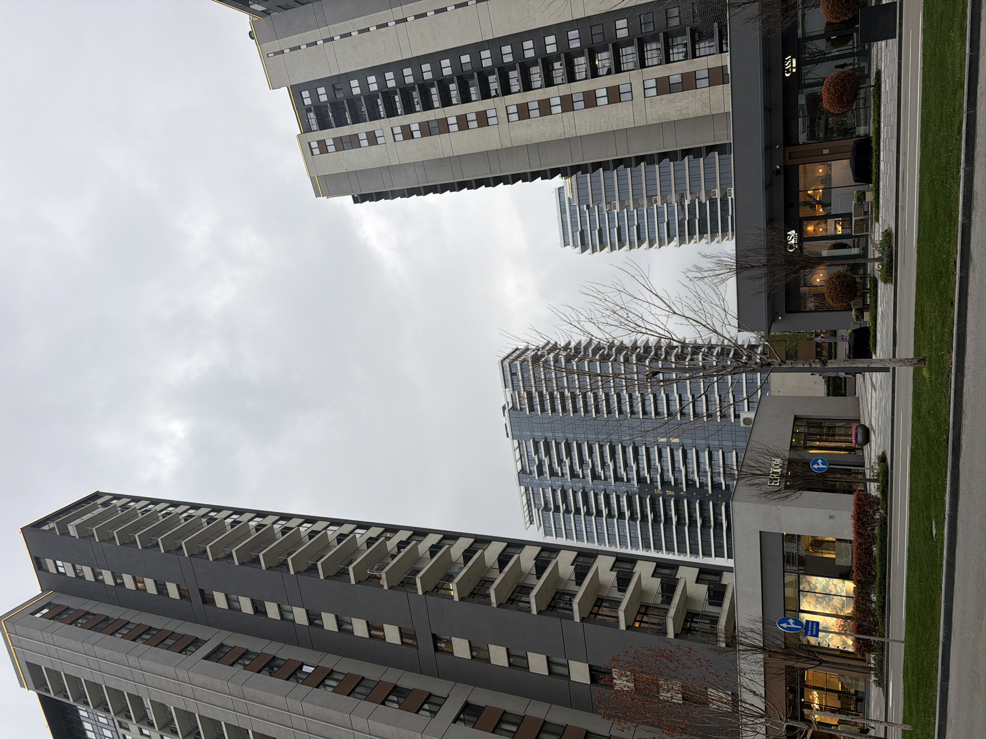 The image shows a view of modern buildings in Belgrade, Serbia, under a cloudy sky. There are three tall residential buildings visible, constructed in a modern architectural style. The buildings have repetitive patterns of windows and balconies. A commercial building is visible in the foreground, with "CASA BY WINS" signage on the left and "EURODOM" on the right. The storefronts are illuminated. Bare trees stand along the street. A paved sidewalk and street are present. A blue traffic sign is visible in front of the Eurodom building. The overall impression is one of a recently developed urban area.