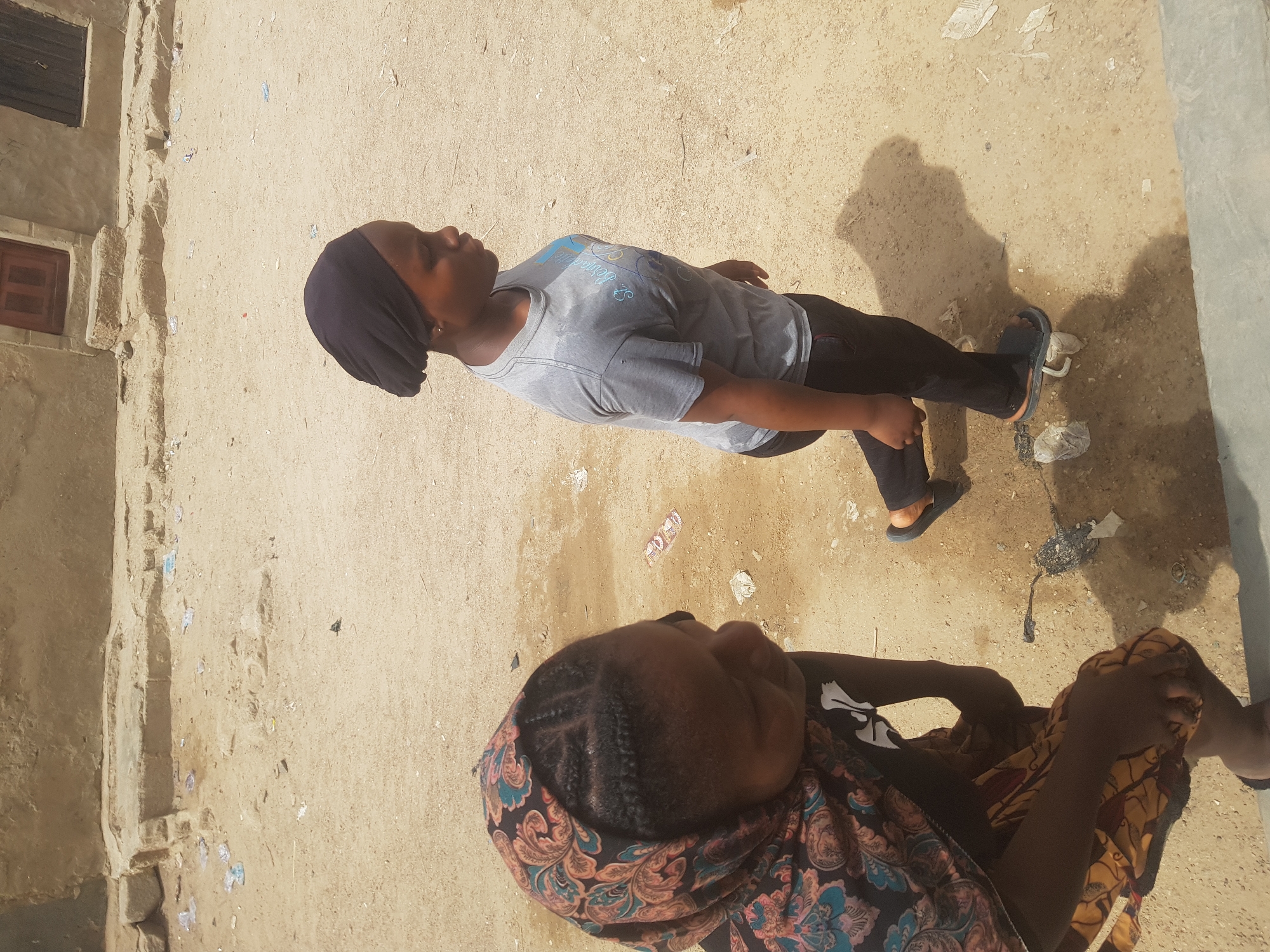 Two individuals are in an outdoor area in Dutsinma A, Nigeria. One person stands wearing a gray t-shirt, black pants, and a black head covering. The second person is seated, wearing a patterned shawl and a black shirt. The ground is sandy and shows signs of debris. A building with a small window is visible on the left side of the frame.