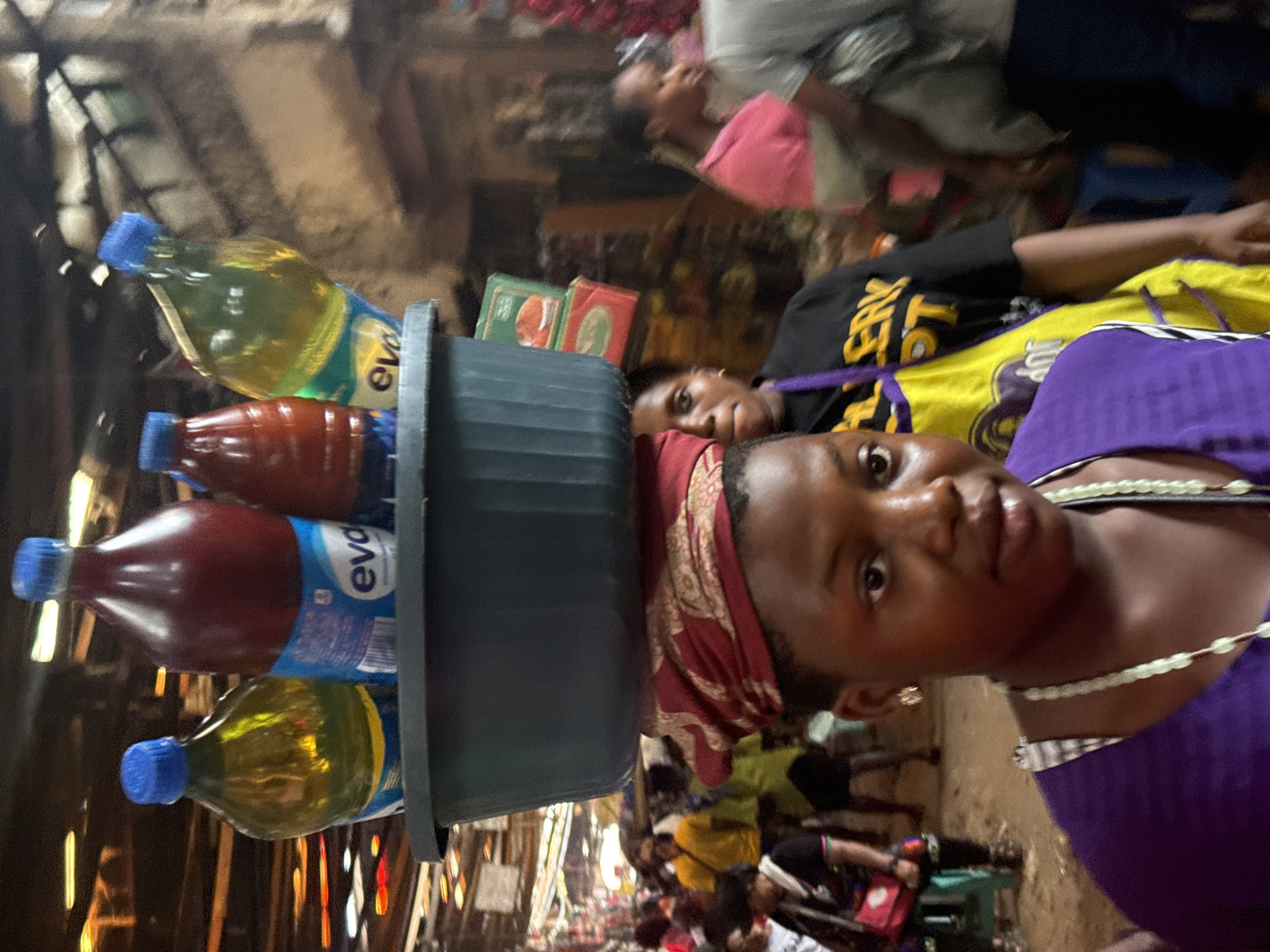 The image depicts a young woman in Onitsha, Nigeria, carrying a gray plastic tub on her head. The tub contains several bottles of Eva-branded beverages. Two bottles appear to contain a yellow liquid, possibly oil, and two bottles contain a red liquid. The woman wears a purple top, a beaded necklace, and a red headscarf. She is looking directly at the camera. The background shows a bustling market scene with people and various goods. Other individuals are visible, including a person in a yellow and black top. The location appears to be a crowded street within the city of Onitsha.