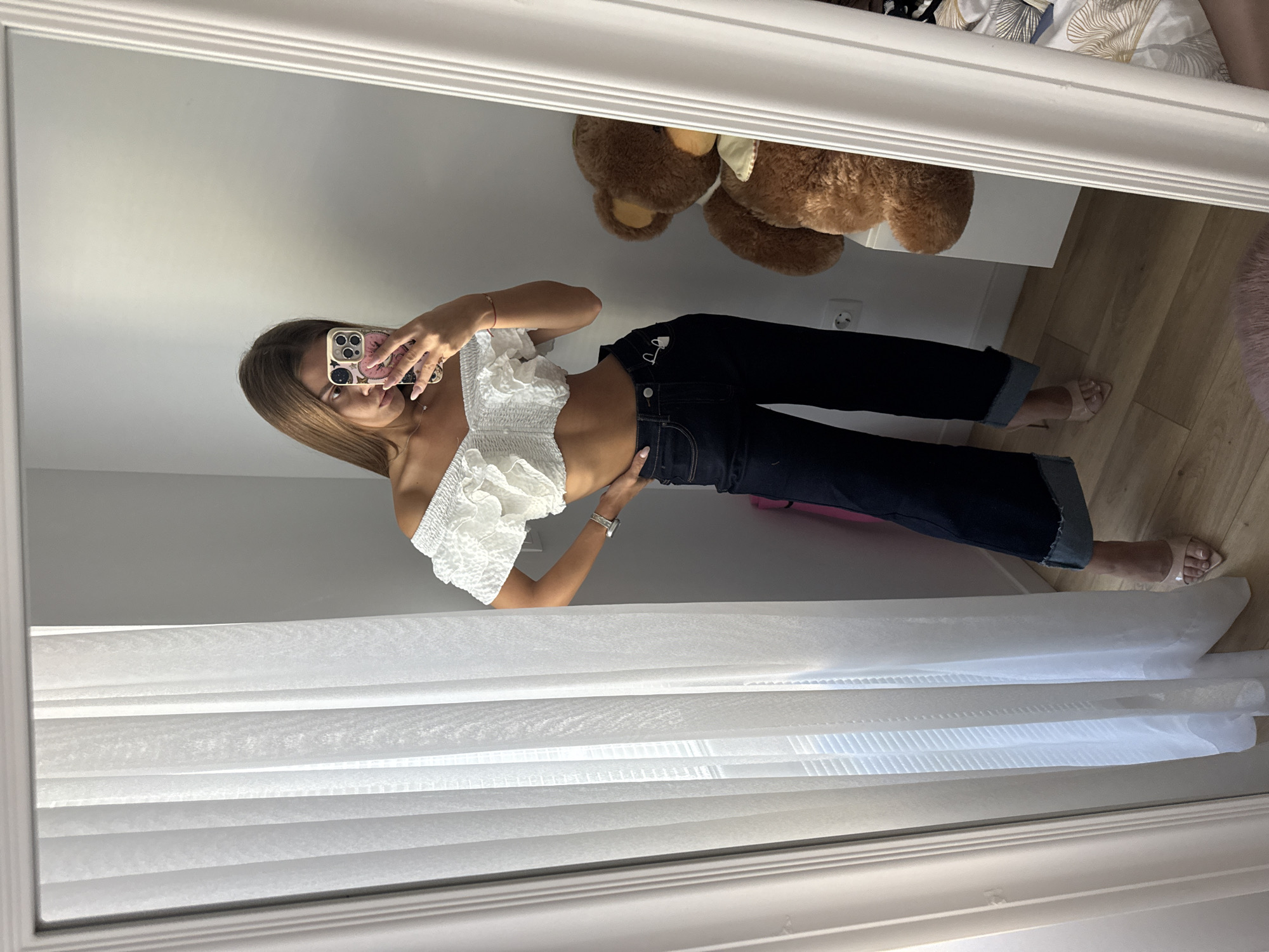 A young woman with long brown hair is taking a mirror selfie in a room. She is wearing a white off-the-shoulder ruffled crop top and dark wash wide-leg jeans with rolled cuffs. She has on nude pointed-toe heeled mules. Her left hand is on her hip, and she is holding a pink phone with a patterned case in her right hand. To her right, a large brown teddy bear sits on a white shelf. The room has white walls, sheer white curtains on the left side, and a light wood floor. The mirror frame is white and ornate. There is a wall outlet visible on the wall behind the teddy bear. The scene does not provide any specific indicators that it is located in the city of Plovdiv, Bulgaria.
