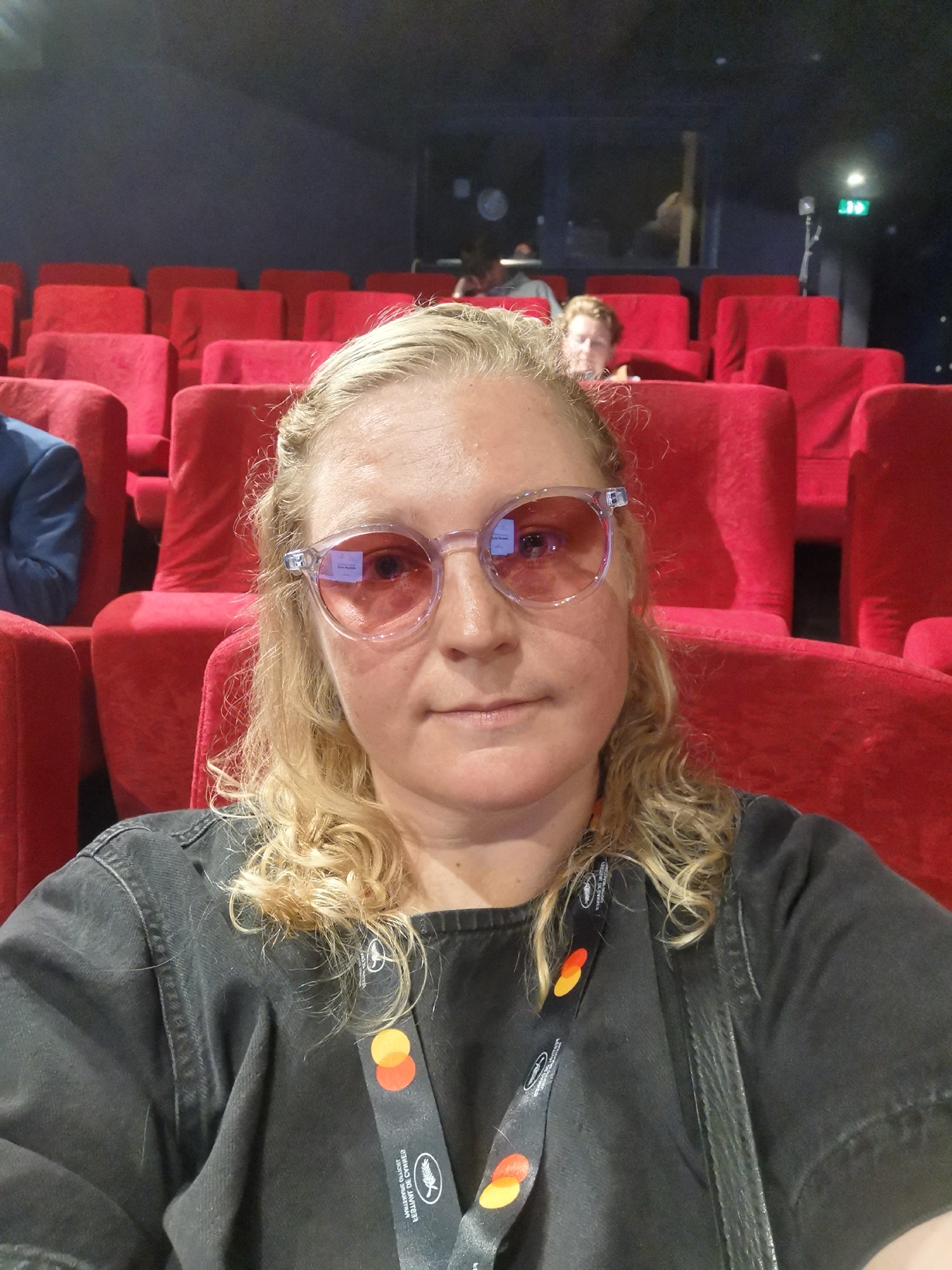 The image shows a blonde woman with curly hair, wearing pink-tinted glasses, a dark-colored top and a lanyard. She is sitting in a red-seated auditorium. There are other people seated behind her, further back in the room. The lanyard around her neck indicates the "Festival de Cannes" branding. This suggests the photo was taken at the Cannes Film Festival, held in Cannes, France. The woman is the primary subject, facing the camera. The background comprises rows of unoccupied red seats and other people in the distance, providing context for the setting.