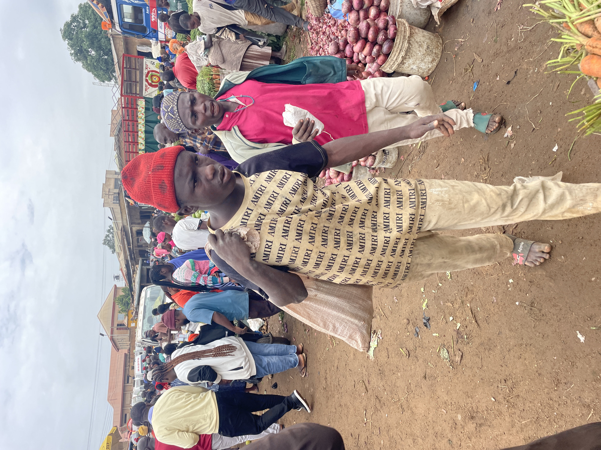 This image depicts a bustling outdoor market scene, likely in Gura Top, Nigeria. Two young men are prominent in the foreground, walking towards the viewer. The man on the left wears a red beanie and a tan t-shirt with repeated black text. He carries a large, light-colored sack over his right shoulder. The man on the right wears a bright pink shirt, a green open jacket, and light-colored pants. He holds a small white packet in his right hand.
Behind them, the market is crowded with people dressed in various attire. Stalls and various goods are visible, including large piles of red onions in buckets, and carrots in a basket in the lower right. Several trucks and vehicles are present in the background, suggesting commercial activity. The ground is a dirt surface, and the sky is overcast. The overall atmosphere is one of a busy marketplace with active commerce.