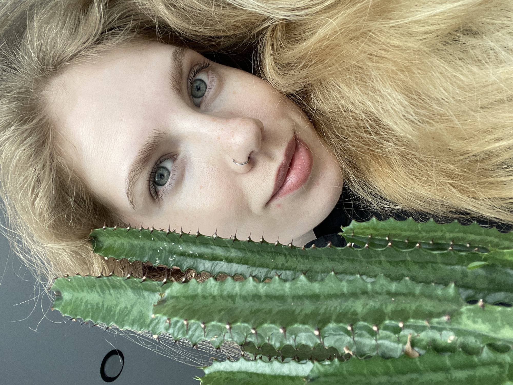 A close-up, tilted view features a fair-skinned person with light blonde, wavy hair partially framing their face. They have light eyebrows, green eyes, and a silver nose ring. Their lips are light pink. The person is partially obscured by a green, multi-segmented cactus with small thorns along the edges. The background consists of a dark, reflective surface. There is no information to confirm this photo was taken in Izhevsk, Russia.