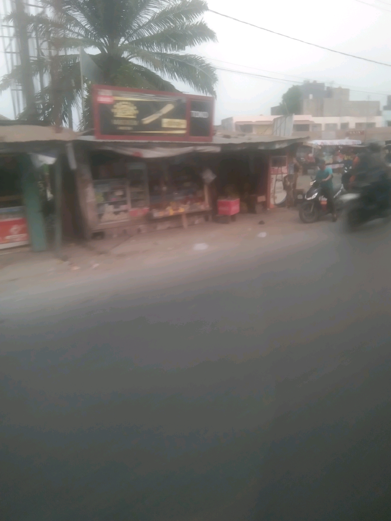 The image shows a street scene with several buildings, some of which appear to be shops, along a road. A palm tree is visible on the left side of the frame, and several motorcycles are parked or in motion on the road. One motorcycle has a person riding it. There are also some structures that appear to be market stalls or small shops. In the background, there are more buildings, some of which are taller. The sky is overcast and the overall lighting suggests it is daytime. The image is somewhat blurry, likely due to motion or low resolution.