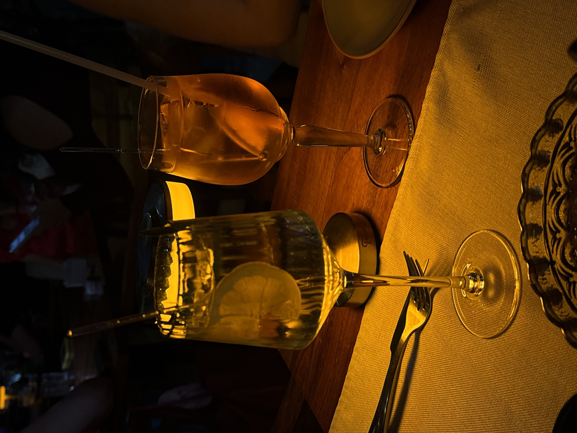 The image displays a dimly lit indoor scene, likely a restaurant or bar, with a warm, golden ambiance. Two stemmed glasses, resembling wine or large cocktail glasses, are positioned on a wooden table. Both glasses contain a light amber or golden-colored beverage, each garnished with a slice of citrus fruit, likely lemon or lime, and a clear drinking straw.
In the foreground, a small light source, possibly a candle or mini lamp, casts a bright golden glow on the base of the nearest glass and the surrounding area. A silver fork is placed on a textured, light yellow or golden placemat next to this glass. To the right, part of a decorative plate or coaster with an intricate scalloped edge and pattern is visible. The second glass is situated slightly further back on the wooden table.
The background is dark and out of focus, with indistinct shapes hinting at other people or elements of the venue. The overall lighting suggests an evening or nighttime setting, creating an intimate and inviting atmosphere. No discernible text is visible within the frame. The original image is oriented vertically, appearing rotated 90 degrees clockwise.