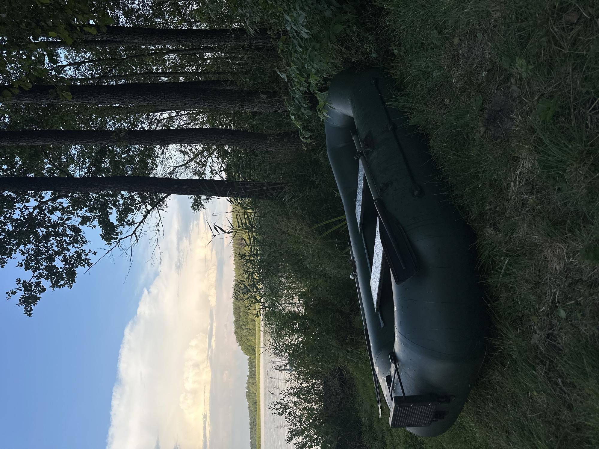 An inflatable, dark green boat rests on a grassy shore beside a body of water. The boat is positioned diagonally in the frame, with its front end facing left and its stern to the right. Oars are visible inside the boat, lying across a wooden slat. In the background, a dense line of trees forms a backdrop, with their dark silhouettes contrasting against a bright, cloudy sky. The water appears calm and reflects the muted tones of the sky and surrounding foliage. The environment suggests a natural, possibly rural, setting. The location is identified as Слабодкаўскі сельскі Савет, Belarus.