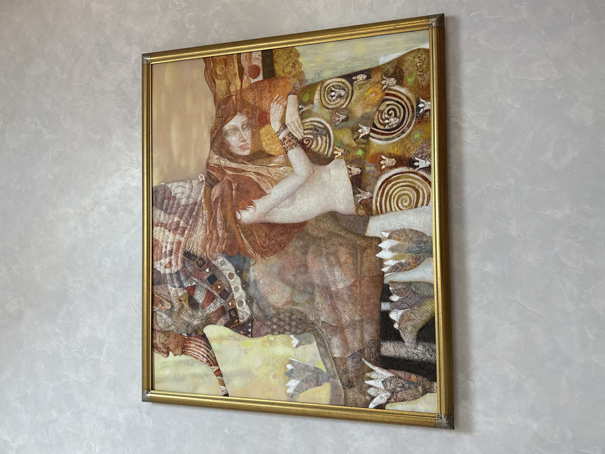 A framed oil painting, likely titled "The Kiss" and by an unknown artist, is displayed on a textured light grey wall. The painting depicts two figures rendered in a detailed, stylized manner. On the left, a figure with a profile view and a long, flowing beard, adorned with a headdress of red, white, and brown patterned fabric, is visible. To the right, a reclining female figure with long, wavy red hair is shown. She is wearing a white garment on her upper body and a richly patterned skirt with floral motifs and swirling designs in shades of gold, green, and brown. The background of the painting is a soft, warm gradient of yellow and orange, suggesting a sunset or ethereal light. Several stylized white and grey floral or plant elements are present at the bottom of the painting. The painting is housed in a thin, ornate gold-colored frame. The setting is an indoor environment, with the wall providing the primary environmental context. The image was captured in Yukki, Russia.