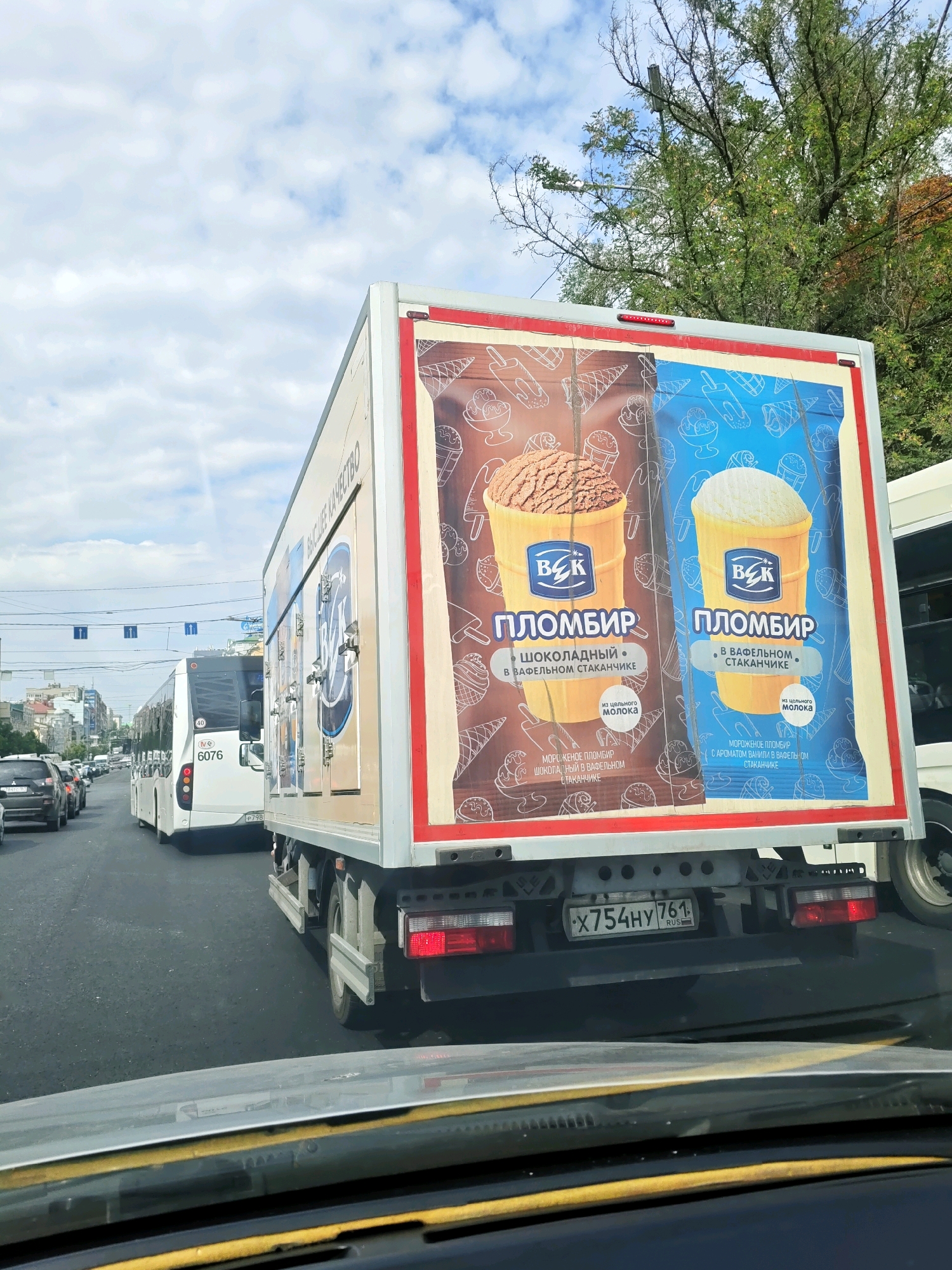 The image shows a street scene in Rostov-on-Don, Russia, likely on a sunny day with a cloudy sky. The foreground is dominated by the interior of a car, with the dashboard and windshield visible. The main subject is a large white truck with colorful advertisements for ice cream on its rear. The advertisements feature images of ice cream cones and cups, with text in Russian. The truck has a license plate that reads "X754HY 761 RUS".
In the background, several other vehicles are visible, including a white bus with the number "6076" on its rear, and other cars. The bus also has a license plate. A street with traffic lights and overhead wires is present. Trees with green foliage line the street. Buildings are visible in the distance. The overall impression is a busy urban environment.
