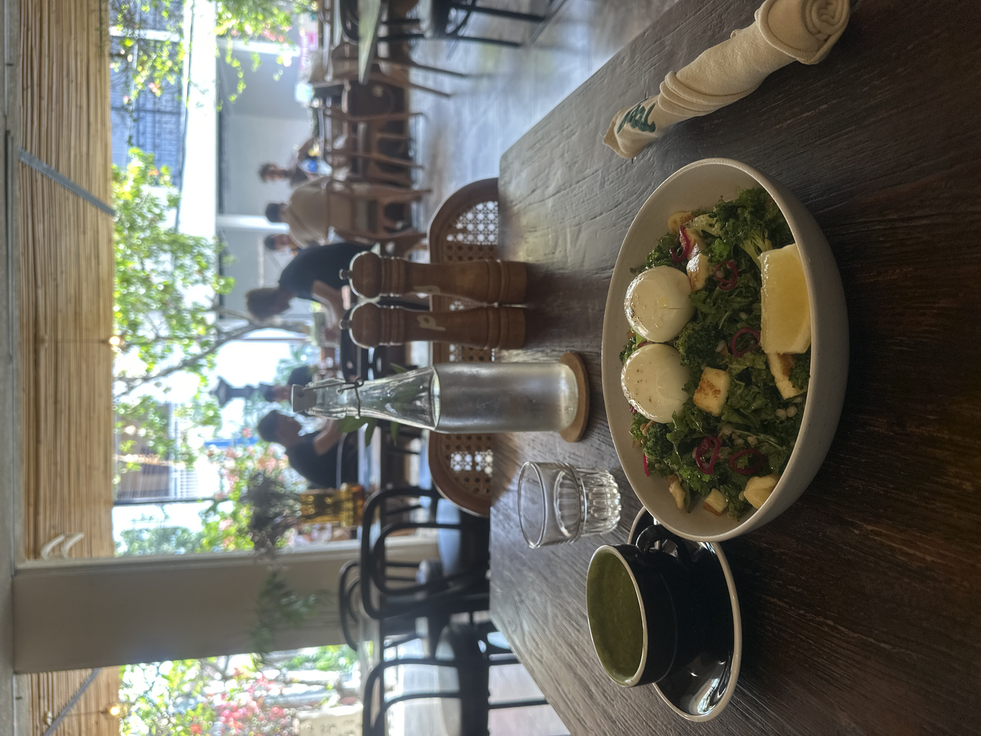 In the foreground, a dark wooden table holds a breakfast salad, a cup of green tea, a glass of water, and a bottle of water with salt and pepper shakers. The salad is in a white bowl and topped with two poached eggs. The cup of green tea is black, and the saucer is also black. The glass of water is clear, and the bottle of water is made of clear glass. In the background, people are seated at tables in a restaurant with bamboo blinds on the windows, allowing natural light to filter in. Plants are visible through the windows, suggesting an outdoor or garden setting. The overall scene depicts a casual dining experience.
