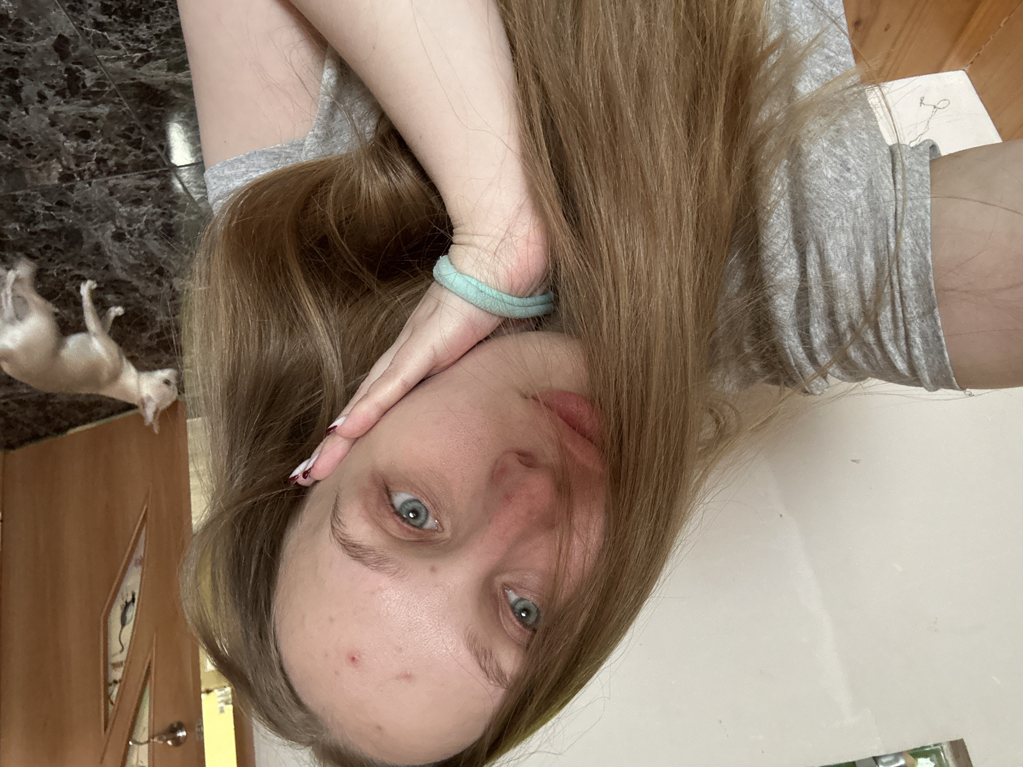 A person with fair skin and long, light brown hair lies with their head angled towards the camera, hand near their face with a light blue hair tie on their wrist. They are wearing a gray t-shirt. The person has blue eyes. In the background, there is a marble-like dark countertop, a wooden door with a decorative panel, and a light-colored dog lying on its back with its paws up. The background also includes a white surface. The location is Chaikovsky, Russia.