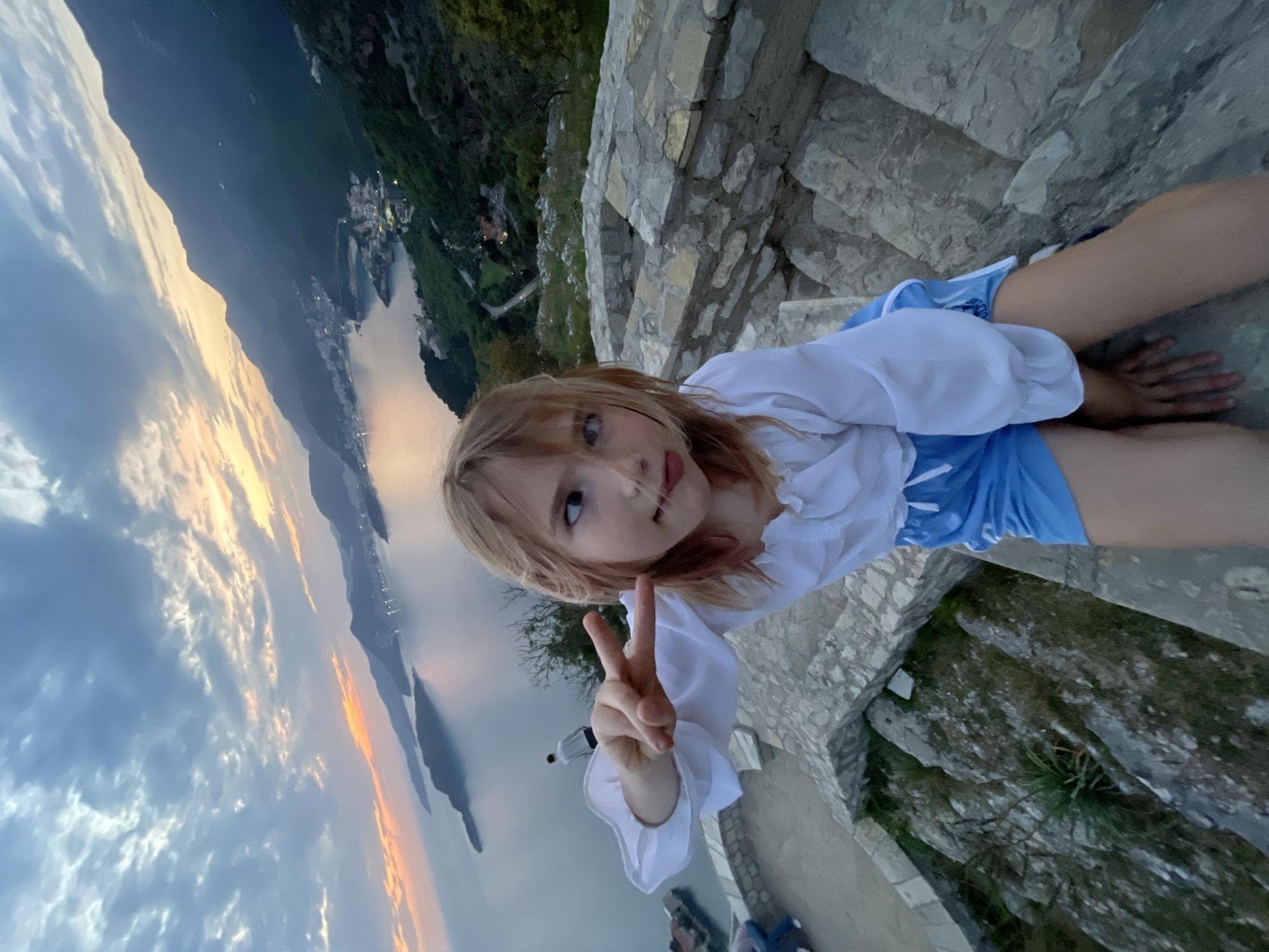 A young girl is featured prominently in this vertically oriented, high-angle shot, likely taken during sunset in Sveti Stefan, Montenegro. She has light-colored hair and is wearing a white ruffled blouse and blue shorts. The girl looks directly at the camera with a slight smile and makes a peace sign with her right hand. She appears to be sitting or kneeling on old, roughly cut stone steps or a wall, indicative of a historical or natural stone structure.
Behind the girl, a stunning coastal panorama unfolds. A vast body of water, possibly a bay or the Adriatic Sea, stretches into the distance, reflecting the warm, soft light of the setting sun. Along the water's edge, a distant settlement begins to twinkle with artificial lights. Towering, tree-covered mountains rise steeply from the coastline, dominating the background. The sky above is a canvas of evolving colors, transitioning from soft blues at the top to vibrant oranges and pinks near the horizon, typical of a beautiful sunset or sunrise. Scattered clouds add texture to the atmospheric sky. The scene conveys a sense of serene natural beauty and the joy of observing it from an elevated viewpoint. No text is visible in the image.