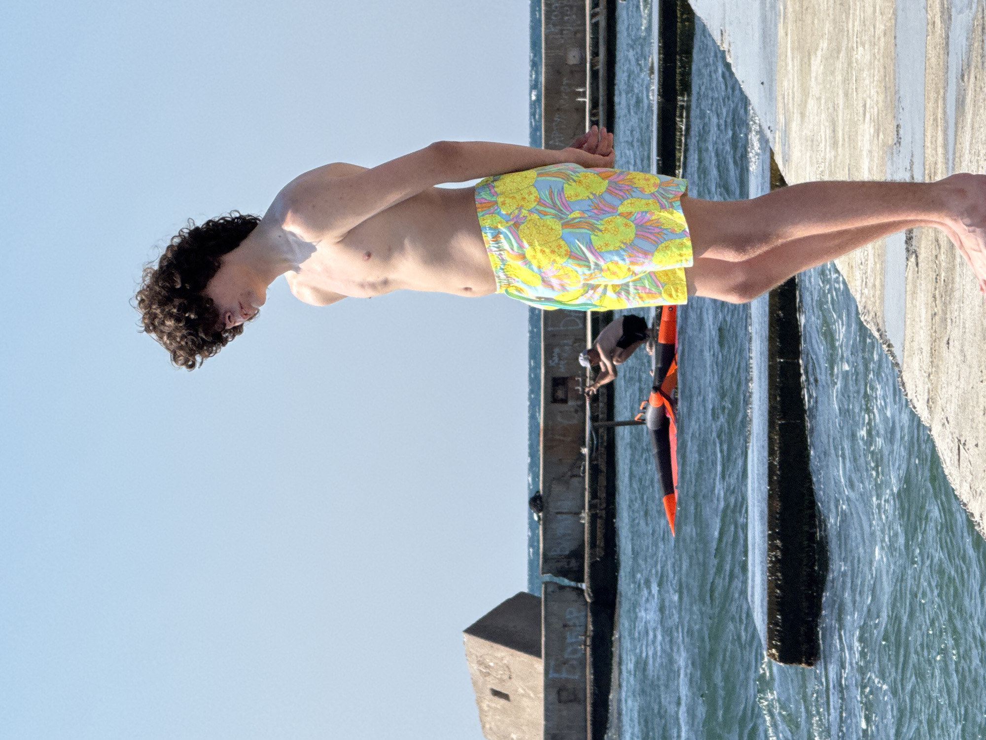 A shirtless young man with curly dark hair stands on a concrete pier, facing away from the camera. He is wearing brightly colored swim trunks with a pineapple pattern. In the background, the sea is visible, with a person on a watercraft in the distance. There are also concrete structures along the coastline, one with graffiti on it. The sky is clear and blue. The image appears to have been taken in Sevastopol, Ukraine, given the context of the user's request.