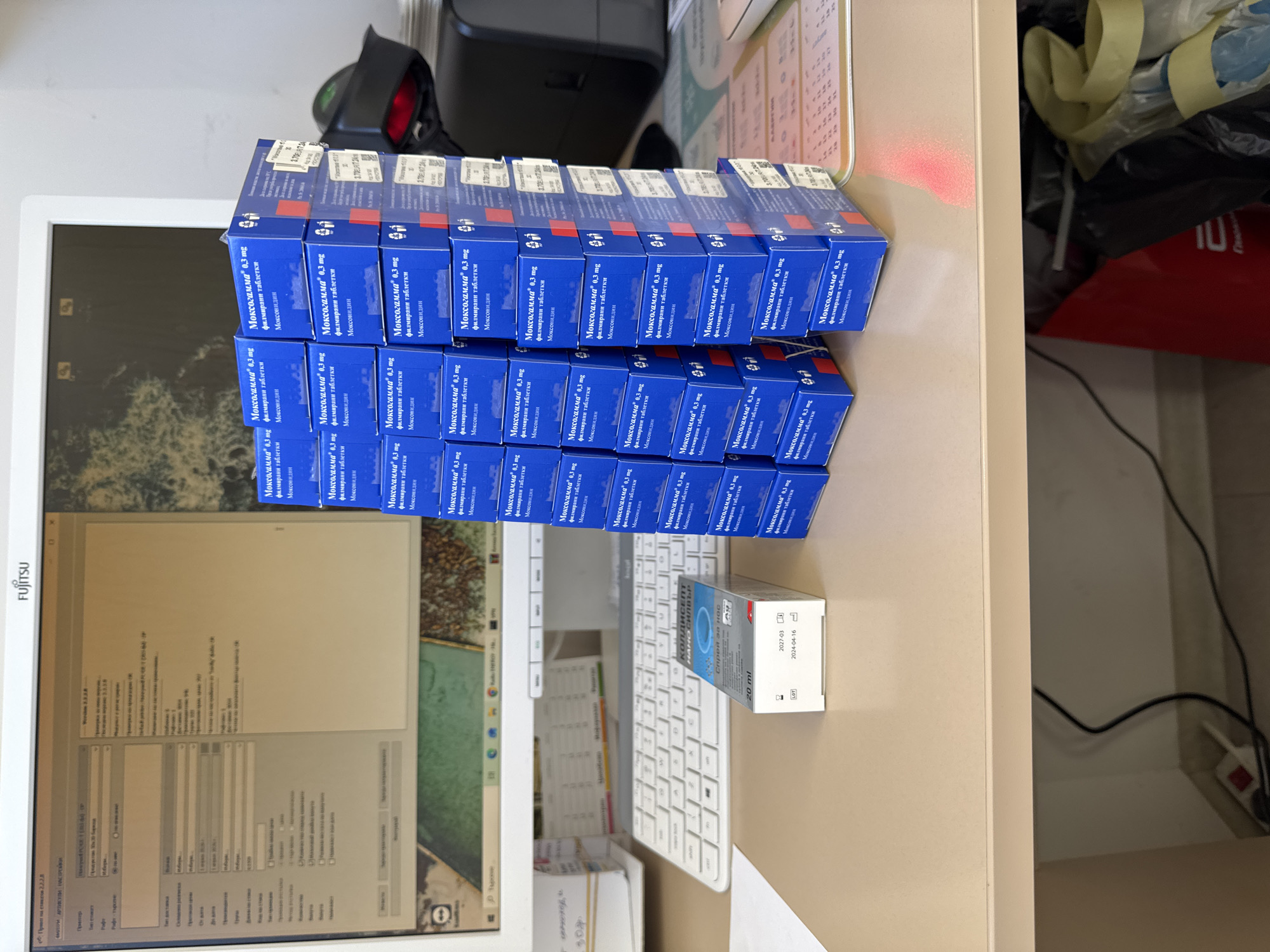 The image shows a desk with a Fujitsu computer monitor displaying a software interface with Bulgarian text, a white keyboard, and a stack of "Moxogamma 0.3 mg" medication boxes. To the right of the keyboard sits a "Koldisept Nano Silver" nasal spray box. A black scanner is behind the medication. The context does not provide information about the city of Rudozem or any people.