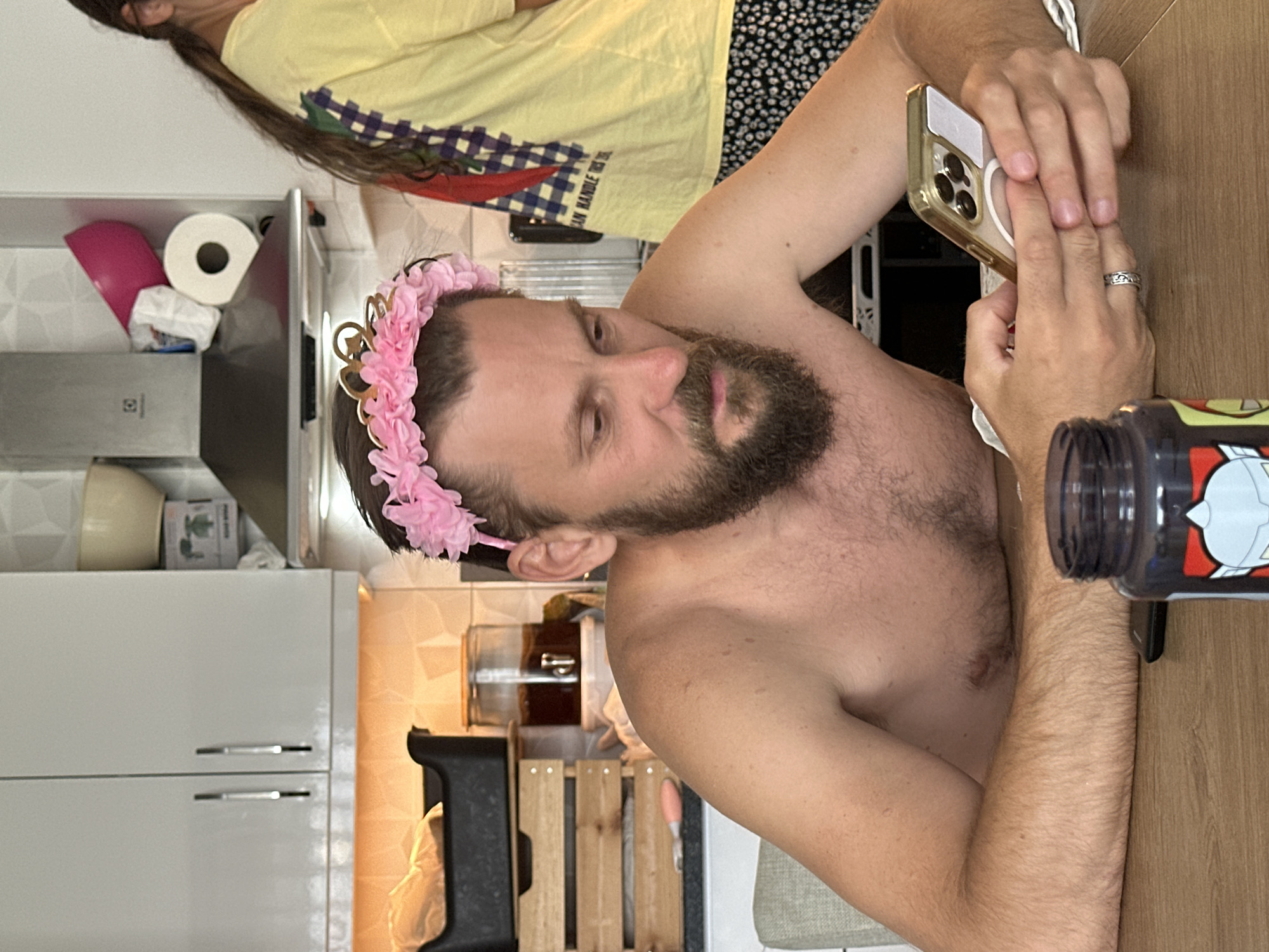 A shirtless man with a beard and a pink flower crown is looking at his phone. He is sitting at a table with a water bottle next to him. A woman wearing a yellow shirt and black and white patterned pants is standing behind him. They are in a kitchen with white cabinets and countertops.