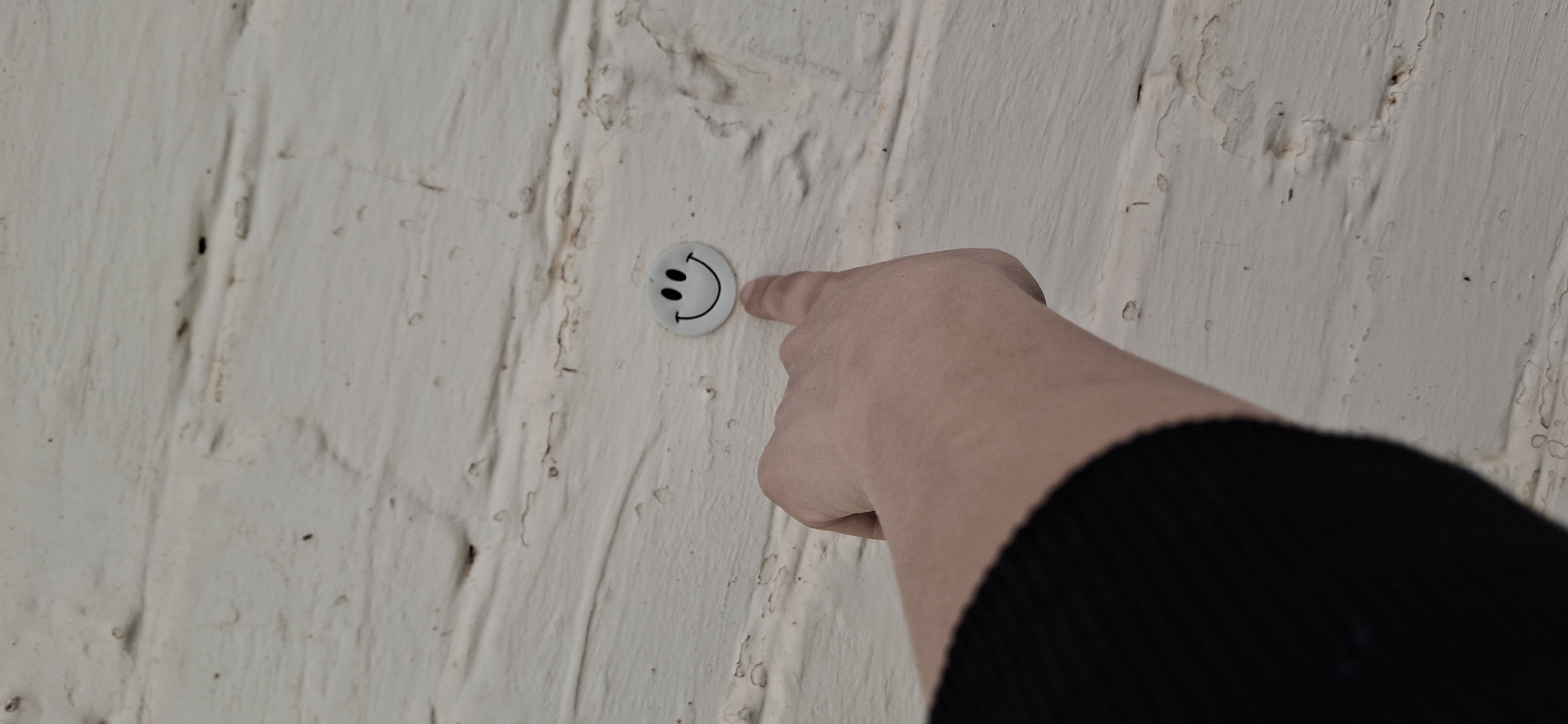 A pale-skinned hand with a finger pointing towards a white circular object with a black smiley face imprinted on it, which is affixed to a textured, light-colored wall, likely an interior brick wall with a rough painted surface. The hand and arm are positioned on the right side of the frame, with the arm clad in a black, knitted sleeve. The background reveals details of the wall's texture and subtle variations in its coloring. The location in the city of Писарёвка, Russia is not discernible from the visual data.