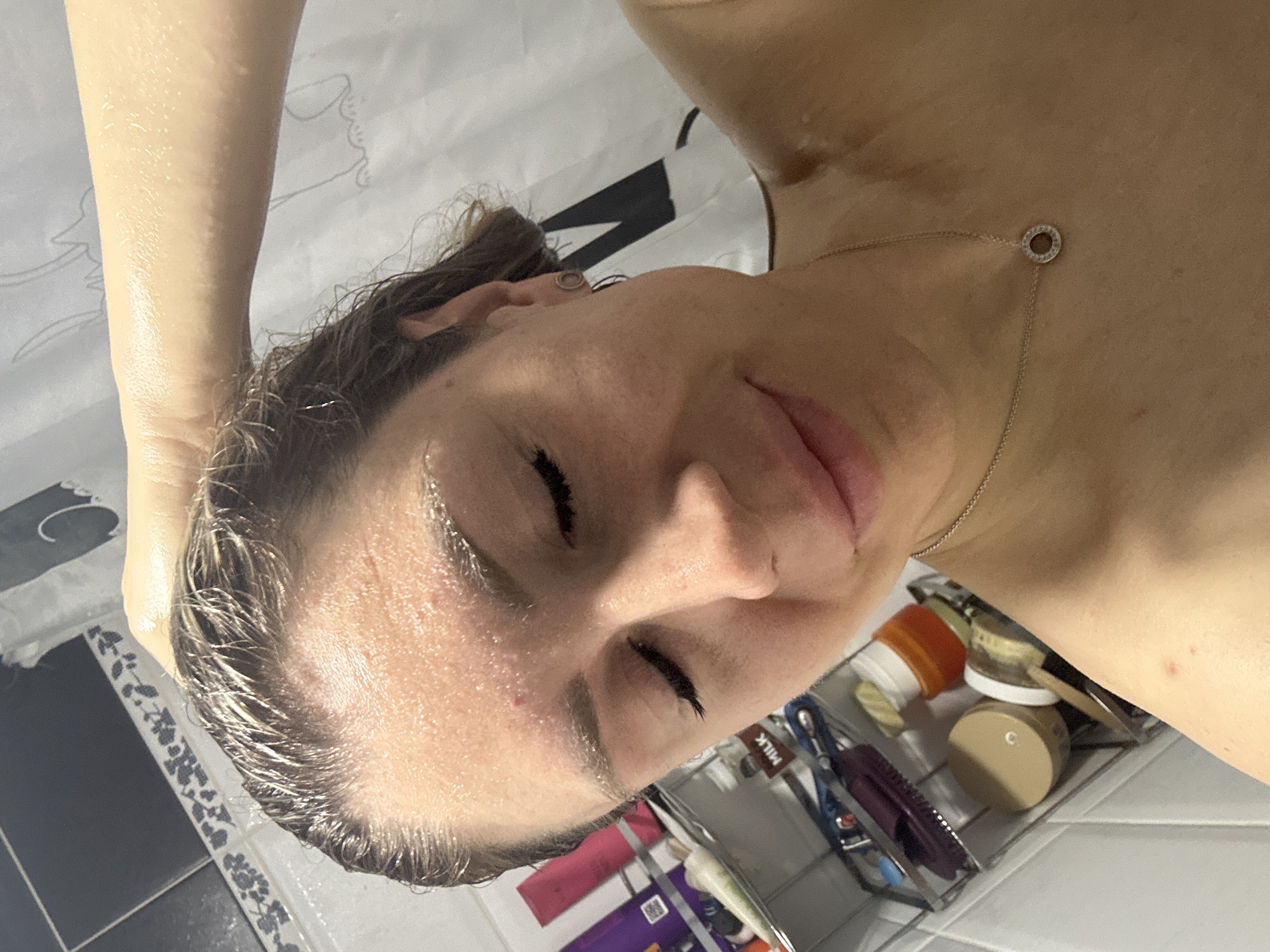 The image shows a close-up, selfie-style shot of a woman in a shower. She has wet hair and is touching her head with her right hand. She is wearing a necklace. Various toiletries are visible on a shelf in the background. The scene appears to be a domestic bathroom. The location in Novoaltaysk, Russia is not discernible from the image.