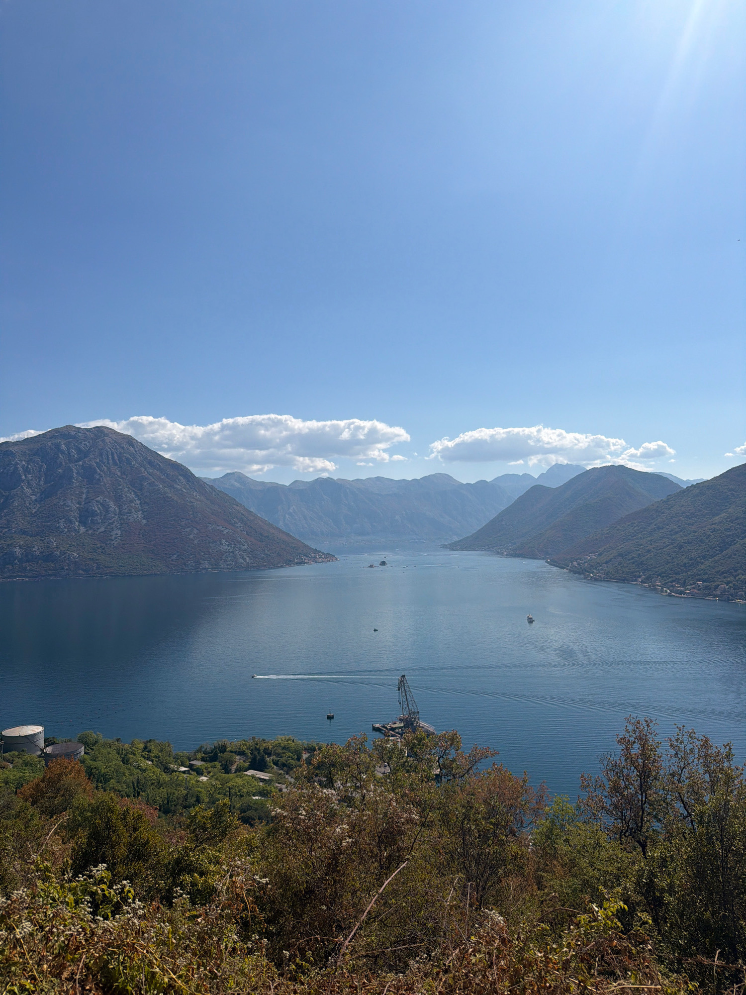 An aerial, full shot captures a wide, deep blue bay embraced by lush, green mountains under a clear, bright blue sky with scattered white clouds. The bay's surface is calm with gentle ripples. A white boat is actively navigating across the bay, leaving a visible wake. In the mid-distance, a prominent industrial crane structure is visible on a floating platform. In the foreground, dense green and brown foliage, including trees and shrubs, dominate the lower portion of the frame. In the far distance, the mountains fade into a hazy blue, suggesting depth and vastness. The scene appears to be a scenic coastal landscape in the area of Morinj, Montenegro, known for its natural beauty and the expansive bay.
