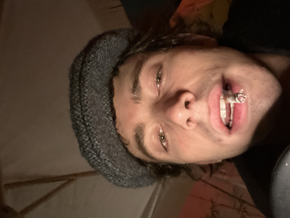 A close-up, low-light image depicts a male individual, appearing to be an adult, from a low-angle perspective. The subject wears a grey, textured flat cap, under which dark, curly hair is visible. His light-colored eyes are directed towards the camera. A lit cigarette or hand-rolled smoking implement is held horizontally between his open lips, with the glowing ember clearly visible. The background is dark and out of focus, revealing indistinct structural elements, potentially part of an overhead canopy or framework, in the upper left quadrant. A faint reddish-orange illumination is present on the left side of the frame. The setting appears to be an indoor or sheltered outdoor area during nighttime. The specific location within Paris, France, cannot be determined from the visual information provided.