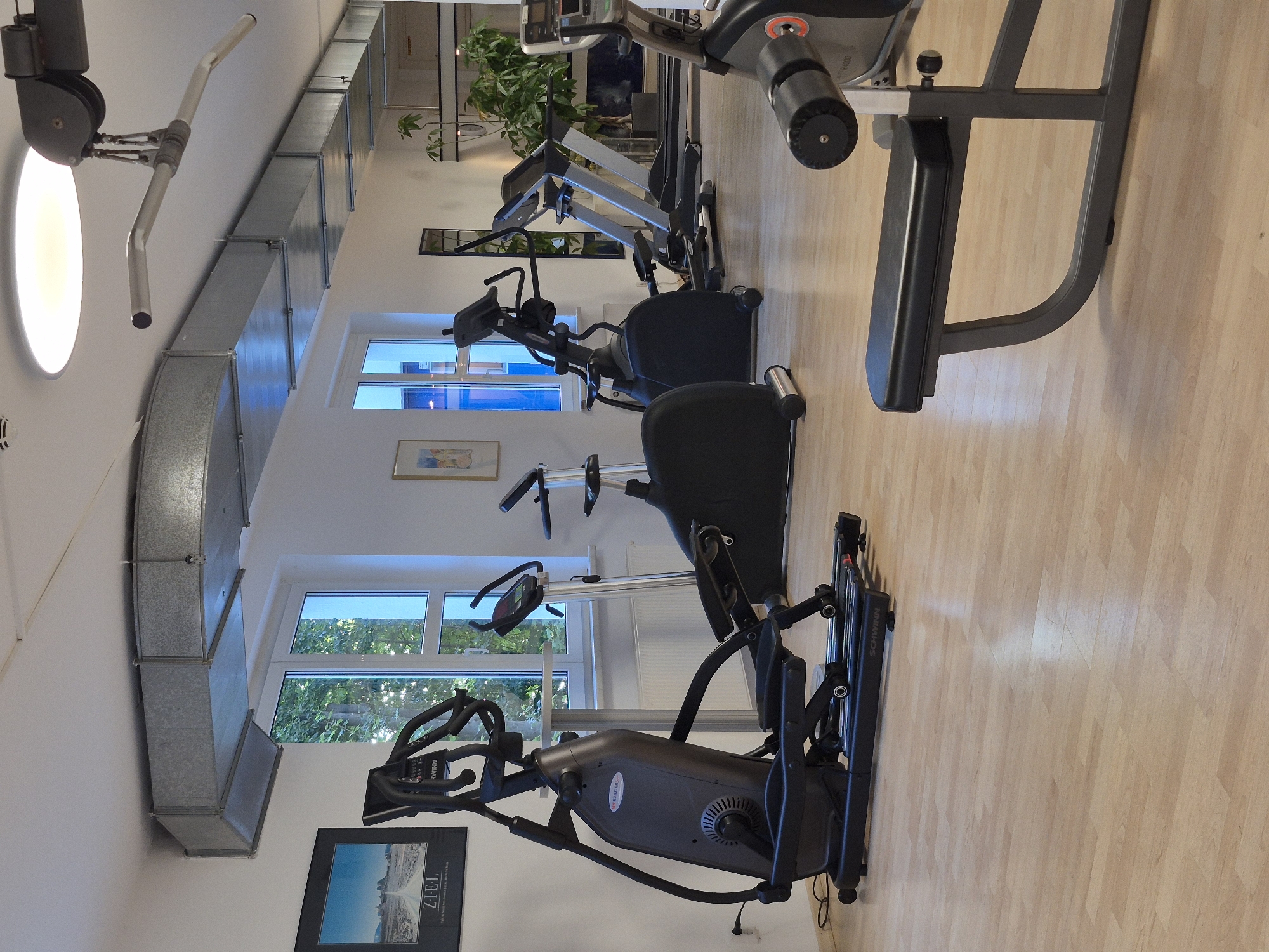The image depicts an indoor fitness facility, likely a gym, located in Schwäbisch Gmünd, Germany. The scene is brightly lit and orderly, with various exercise machines arranged on a light-colored wooden floor. Prominently featured are several cardio machines, including an elliptical trainer on the left, an upright stationary bike, and at least two treadmills in the background. A weight bench is visible in the foreground to the right. The room has large windows that allow natural light to enter, and one wall displays a framed motivational poster with the word "ZIEL" (German for "goal"). The ceiling features industrial-looking ductwork and a circular light fixture. No individuals are present in the image.