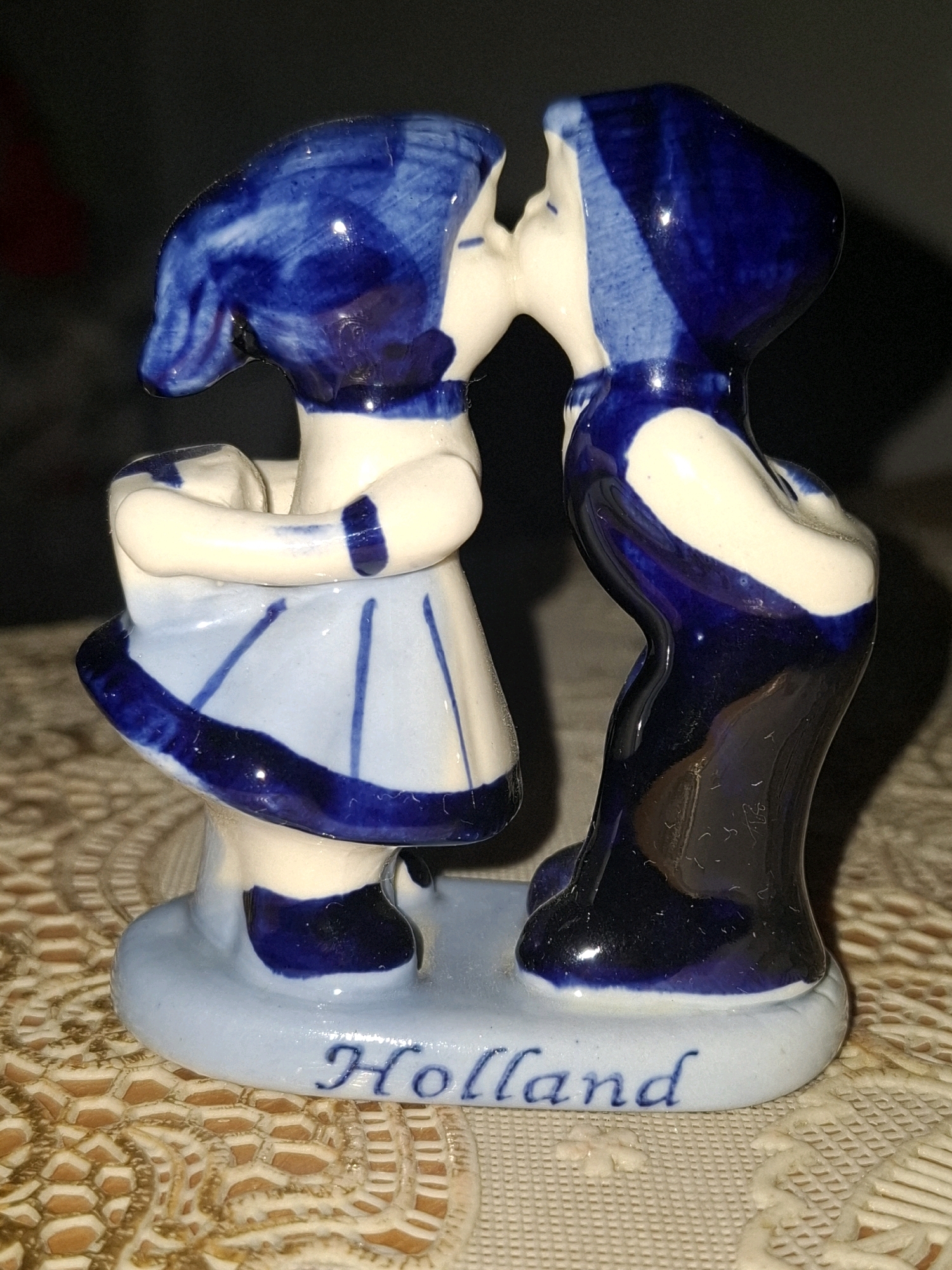 A ceramic figurine depicts two young figures, a boy and a girl, in a tender embrace and about to kiss. They are both adorned in blue and white clothing, with the girl wearing a ruffled skirt and the boy a dark blue garment. The figurine is set on a light blue base inscribed with the word "Holland" in blue cursive. The background is dark and out of focus, suggesting an indoor setting with artificial lighting. The scene conveys a sense of youthful innocence and affection. The figurine's style suggests it might be a souvenir or decorative item, possibly representing a Dutch cultural motif. The visible text "Holland" explicitly identifies a possible origin or theme of the piece. The overall lighting is somewhat dim, with highlights reflecting off the glossy ceramic surface. There are no indications of time of day or weather conditions. The texture of the surface beneath the figurine is visible and appears to be a patterned tablecloth or placemat.