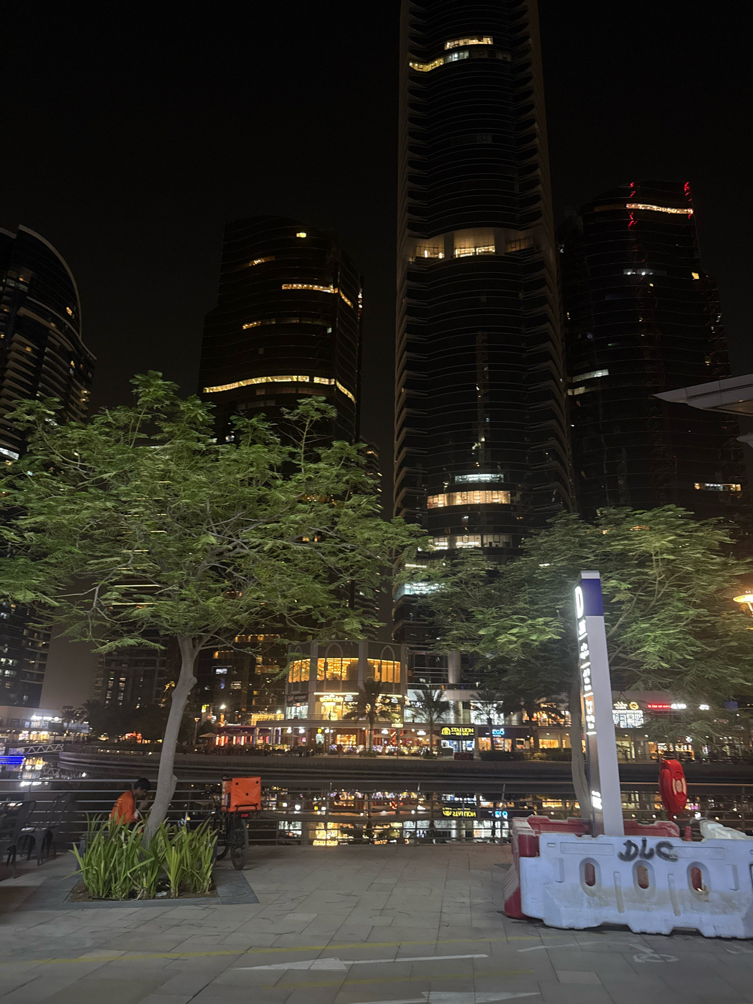 The image depicts a nighttime scene in Dubai, United Arab Emirates. In the background, several tall, modern skyscrapers with illuminated windows dominate the skyline. The buildings have a sleek, contemporary design, with some featuring curved facades. In the middle ground, a body of water reflects the lights of the buildings and businesses lining the waterfront. A tree with lush green foliage is positioned in the foreground, partially obscuring the view of the buildings. The waterfront area appears to be a commercial or entertainment district, with visible storefronts and signs. In the foreground, a paved walkway with yellow markings is visible. There are several objects on the walkway, including a bicycle with an orange delivery box attached, a person wearing an orange jacket and dark pants, and several outdoor chairs and tables. A white and red plastic barrier with graffiti is also present. A street sign with blue and white markings stands near the barrier. The overall atmosphere is that of a vibrant, urban environment at night.
