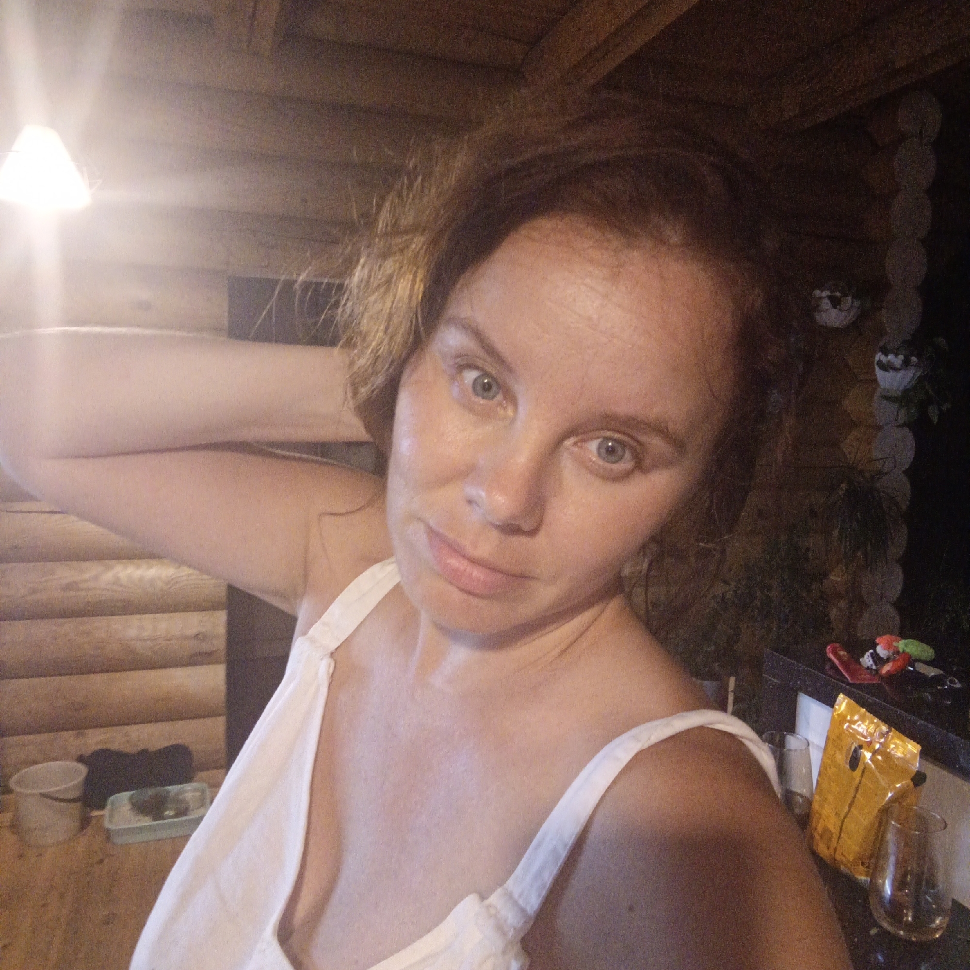 A medium shot selfie is taken of a woman with light brown hair and blue eyes. She is wearing a white sleeveless top. Her left arm is raised and her hand is resting on the back of her head. The background consists of a wooden wall, a ceiling with exposed beams, and some objects on a counter. The scene appears to be indoors, possibly in a cabin or a rustic dwelling. The lighting is warm and somewhat dim, with a bright light fixture visible in the upper left corner. There are also some decorative items and a bag of snacks on a counter in the background. The woman is looking directly at the camera with a neutral expression. The location is identified as Большой Букор, Russia.
