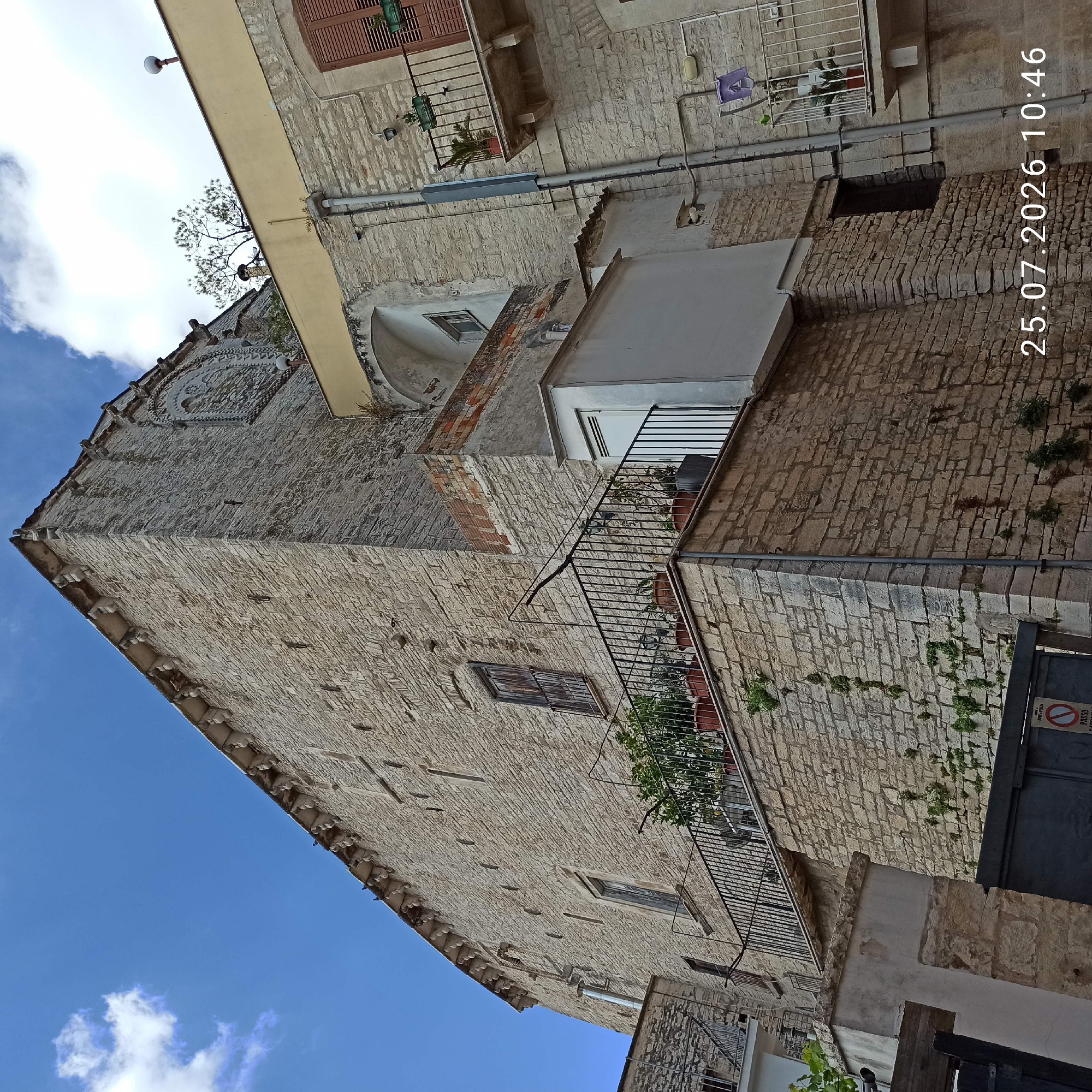 The image depicts an exterior view of an old stone building in Ruvo di Puglia, Italy, with a blue sky and scattered clouds overhead. The building features a large, textured stone facade with visible mortar lines. Several architectural details are present, including a decorative arched window with intricate stonework at the top of the main facade, and smaller, recessed windows along the side.
A metal balcony, adorned with potted plants, extends from one of the upper levels. The balcony has a black metal railing. Below this balcony, another section of the building features more stone construction and a dark metal gate. A smaller building with a white facade and a modern-looking door is attached to the side of the main stone structure.
The scene is viewed from a low angle, emphasizing the height and scale of the stone building. The overall impression is of a historic structure in a European town. A timestamp "25.07.2026 10:46" is visible in the lower right corner of the image. There are no people visible in the frame.
