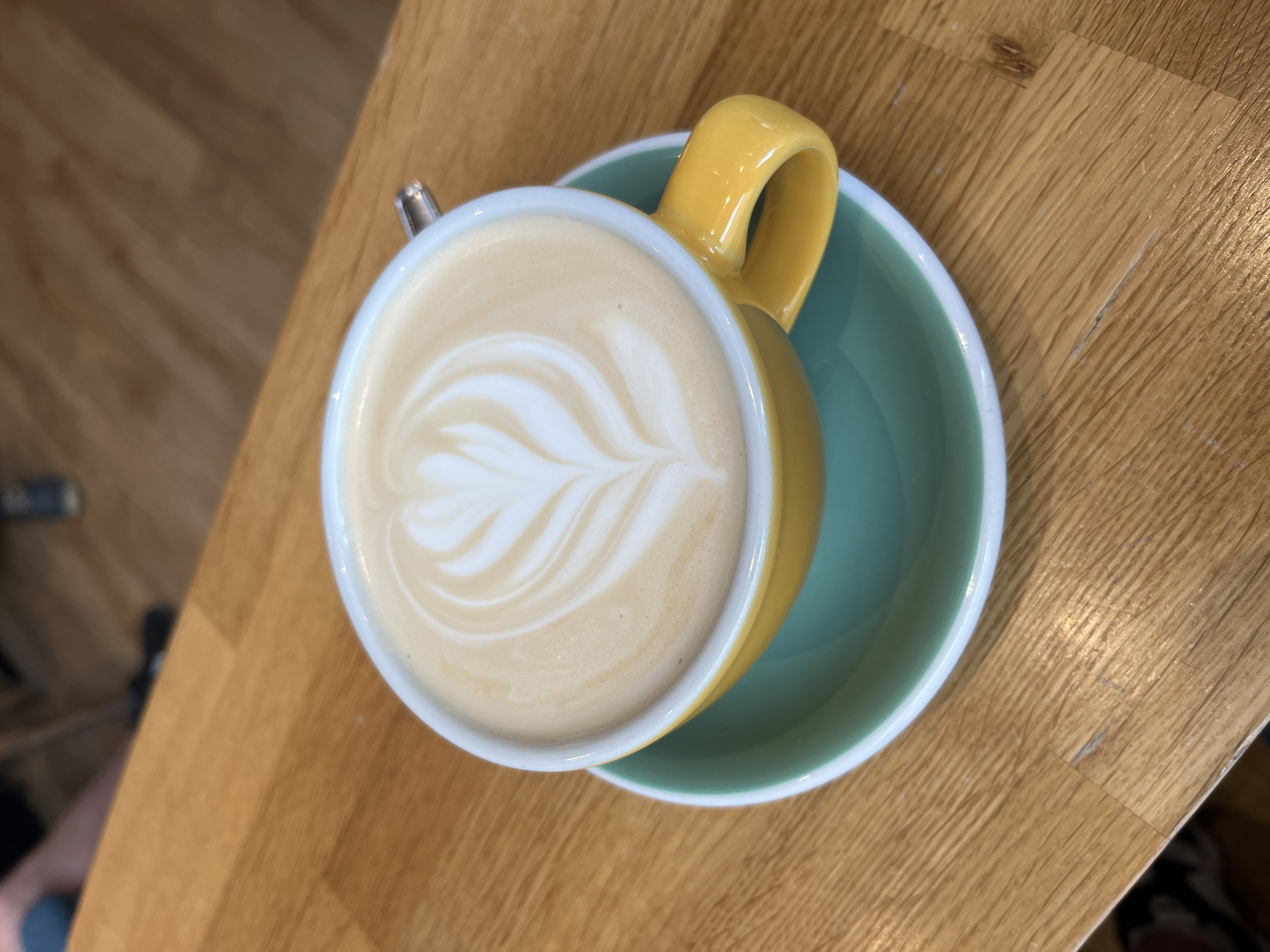 A close-up shot captures a latte with intricate latte art, a yellow mug, and a teal saucer, all resting on a wooden table. The background is blurred, showing glimpses of people's legs and footwear, suggesting a public setting like a café. The lighting is natural, highlighting the creamy texture of the latte and the glossy finish of the mug and saucer. The scene is devoid of any specific landmarks or identifying features that would place it in Pilsen, Czechia, or any other specific city. The focus is solely on the beverage and its presentation.