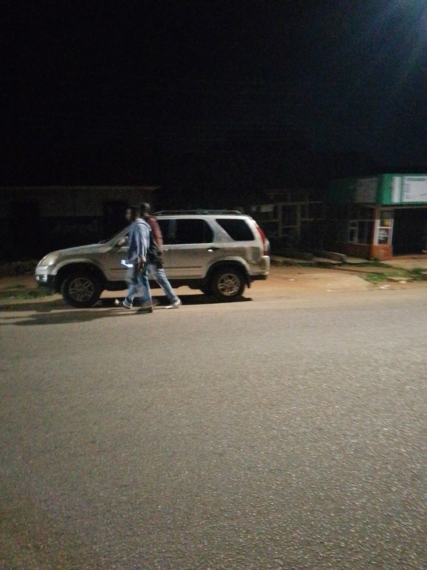Two men walk in front of a silver SUV at night on a paved road. The man on the left is wearing a blue jacket and blue jeans. The man on the right is wearing a dark shirt and blue jeans. The SUV is parked on the right side of the road, facing left. In the background, there is a building with a green sign. The scene is dimly lit, suggesting it is nighttime. The road is in focus, and the people and the car are slightly blurred due to movement or low light conditions.