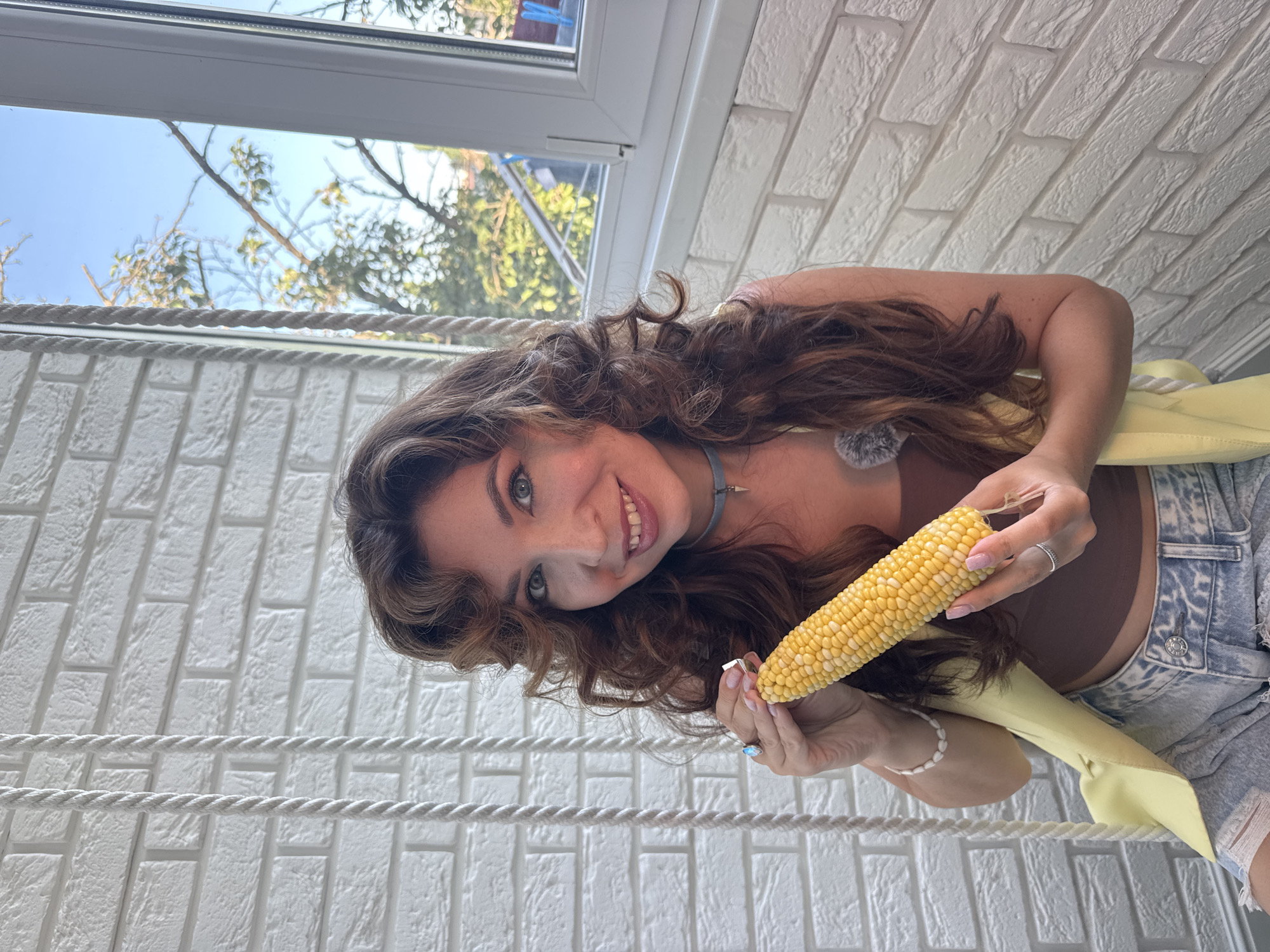 A woman with long, curly brown hair and blue eyes sits on a swing, holding a cob of corn. She is wearing a brown crop top, a yellow vest, and ripped denim shorts. The background consists of a white brick wall and a window with trees and a blue sky visible outside. The scene appears to be outdoors, possibly on a balcony or patio. The location is the city of Kryvyi Rih, Ukraine.