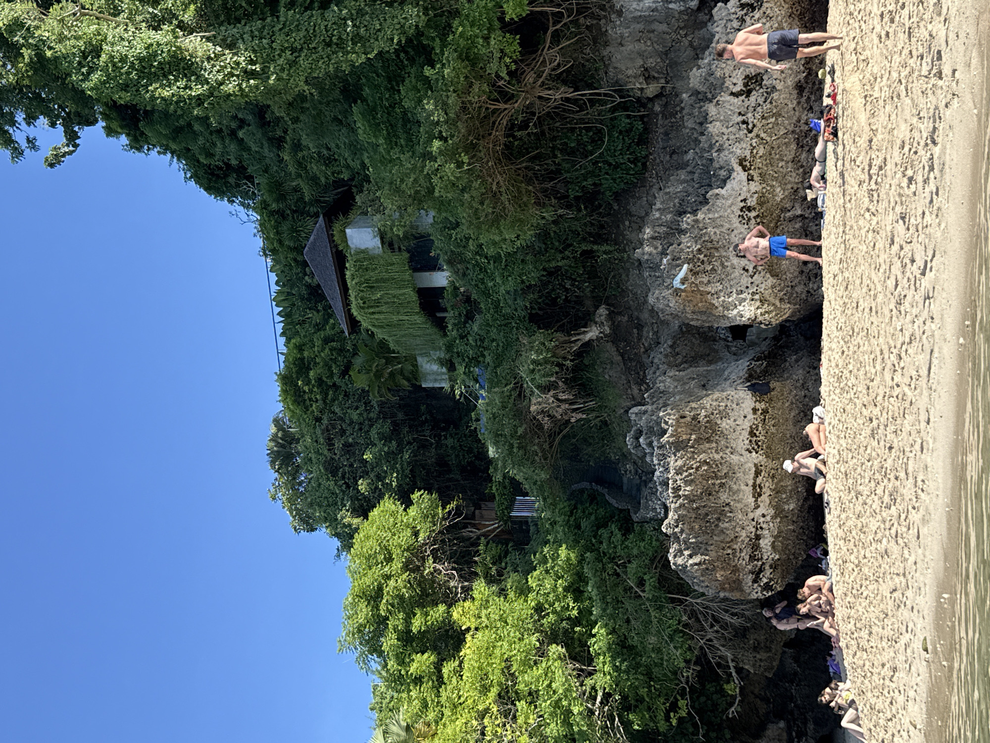 The image depicts a beach scene in Kuta Selatan, Indonesia, with a cliffside partially obscured by dense green foliage. A multi-story building with a dark-colored roof and vine-covered walls is visible amidst the greenery. The lower portion of the image reveals a sandy beach area where people are situated. Some individuals are standing near the rocky cliff face, while others are resting or socializing on the sand. Two men stand near the top of the cliff with only the back of one man visible. Another man is standing along the edge of the cliff wearing blue swimming trunks. The environment suggests a tropical setting.