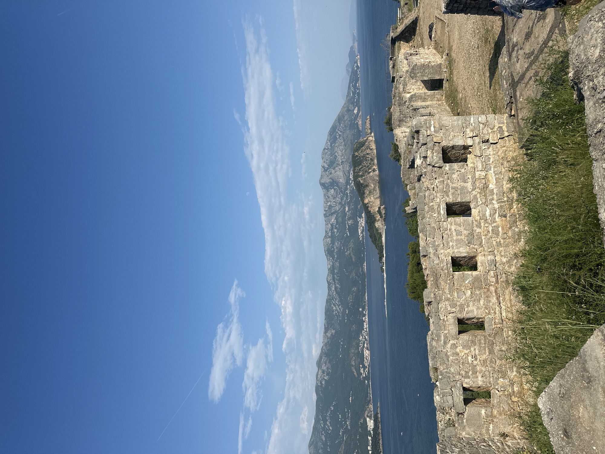 The image depicts a view from the Citadel of Budva, Montenegro. In the foreground, a section of the ancient stone walls of the citadel is visible. The wall features several rectangular openings, likely windows or embrasures. Grass and other vegetation are growing near the base of the wall.
Beyond the wall, the Adriatic Sea stretches out, with a mountainous coastline in the distance. The sky is mostly clear blue with some scattered white clouds. A thin contrail from an airplane is visible against the blue sky. The lighting suggests it is daytime, and the overall setting is a coastal landscape.