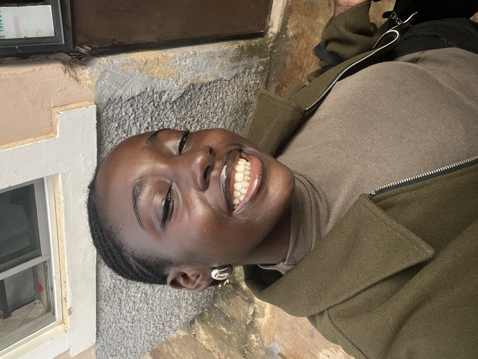 A young Black woman with braided hair is photographed from a high angle, smiling with her eyes closed. She is wearing a dark olive green jacket over a brown turtleneck shirt. Her right ear is adorned with a large silver hoop earring. The background consists of a textured, grayish wall and a window with a white frame, partially obscured by a pinkish-beige colored wall. The scene appears to be outdoors, possibly in an urban environment in Abuja, Nigeria, given the architectural style and general ambiance.
