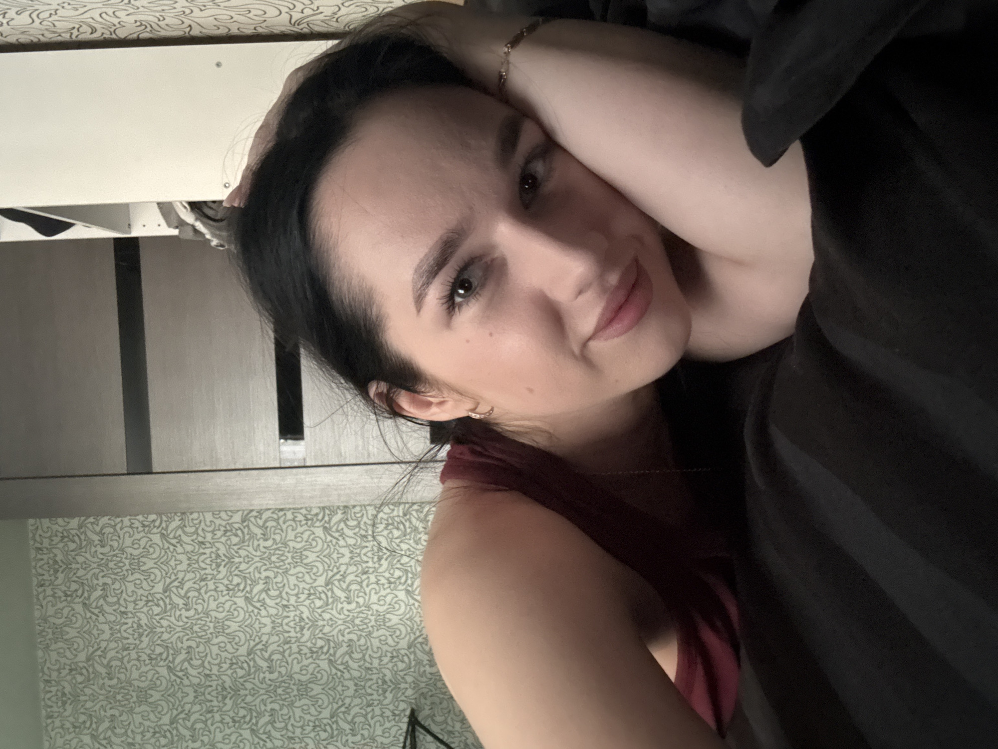 This is a close-up, portrait-style photograph of a young woman lying down. She is resting her head on her arm, which is bent and supporting her head. Her hair is dark and falls around her face and shoulders. She is wearing a maroon-colored top. The background appears to be indoors, with a patterned wallpaper visible on the left side of the image and a piece of furniture, possibly a shelf or cabinet, above her head. The lighting suggests it might be daytime, but the indoor setting and the subdued lighting give a sense of privacy or a casual moment. The woman's expression is relaxed and serene, with a slight smile. She is wearing a delicate bracelet on her arm. There is no visible text in the image. The context provided suggests the location is Novosibirsk, Russia.