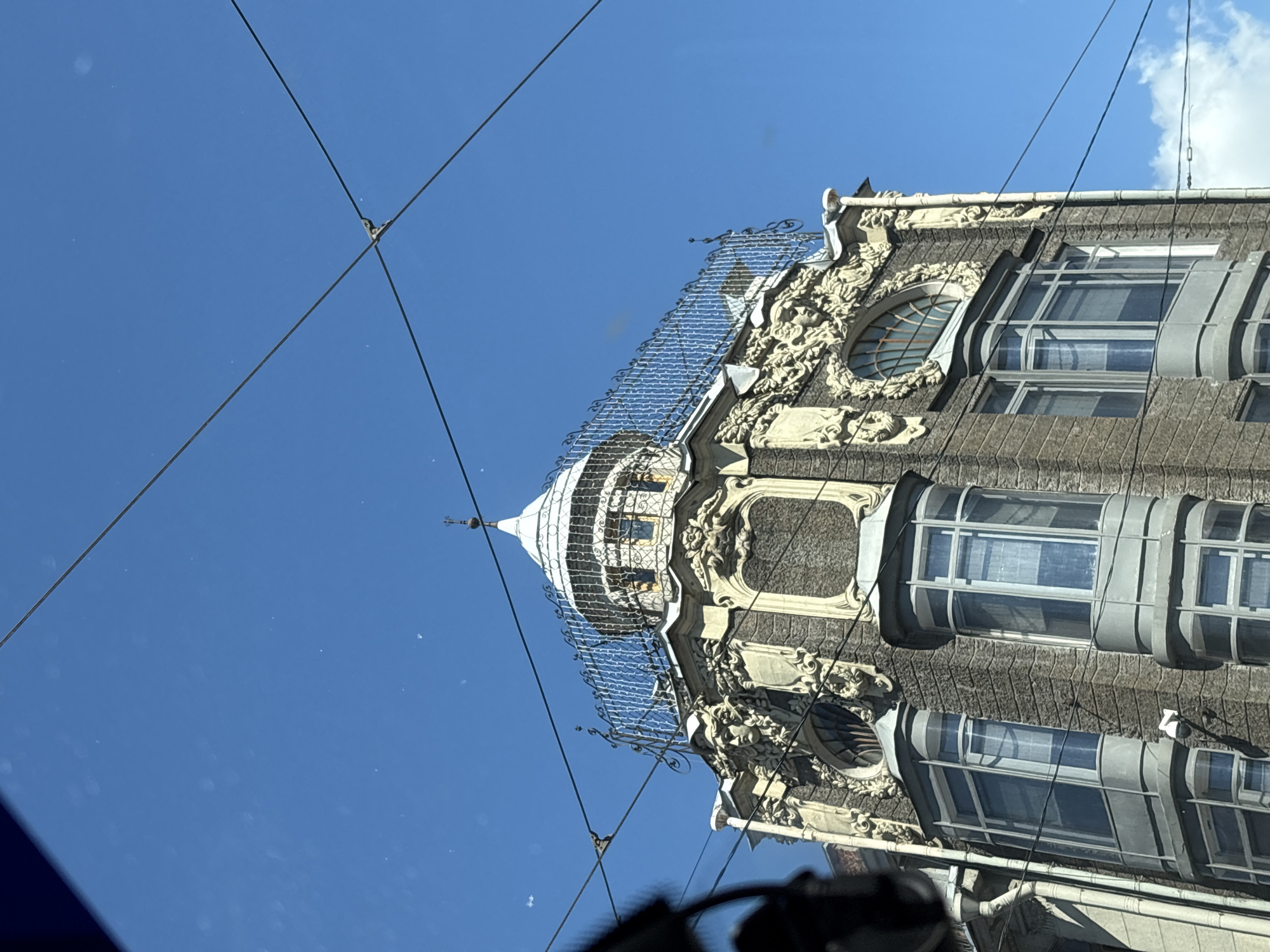 The image shows the upper floors of a building in St. Petersburg, Russia, against a clear blue sky. The building features ornate architectural details, including carvings of faces and floral motifs, bay windows, and a domed turret topped with a decorative spire. Wires, likely for streetcars or telecommunications, crisscross the frame in the foreground and over the building. The building's facade is a mix of stone and decorative elements, with a textured gray stone base and lighter colored ornate embellishments. The overall scene suggests an urban environment on a sunny day.