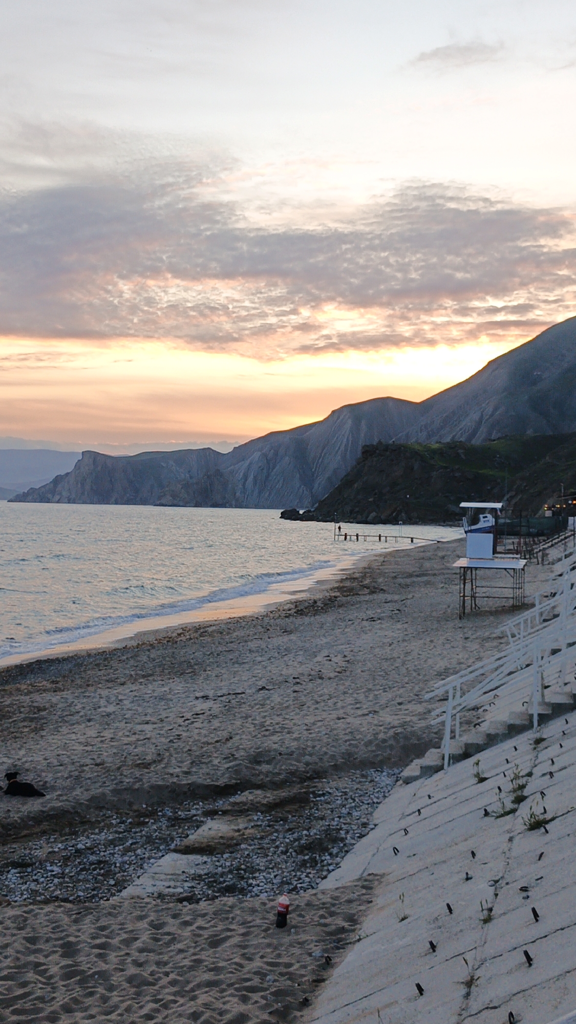 The image shows a beach at sunset in Unknown, Unknown. The beach is sandy and pebbly, with concrete steps and railings leading down to it. In the distance, mountains rise along the coastline. There is a lifeguard stand near the right side. A dog sits in the sand. A soda bottle is lying on the sand near the wall. The sky is filled with clouds that are colored by the setting sun. There are figures on the beach. A pier stretches into the water.