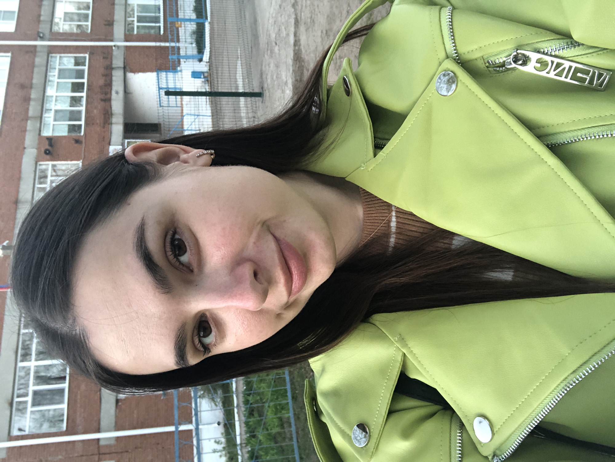 A young woman is posing for a selfie. She has dark hair and is wearing a light green jacket. Behind her is a brick building with white-framed windows and a blue fence. The city is Noviy, Russia.