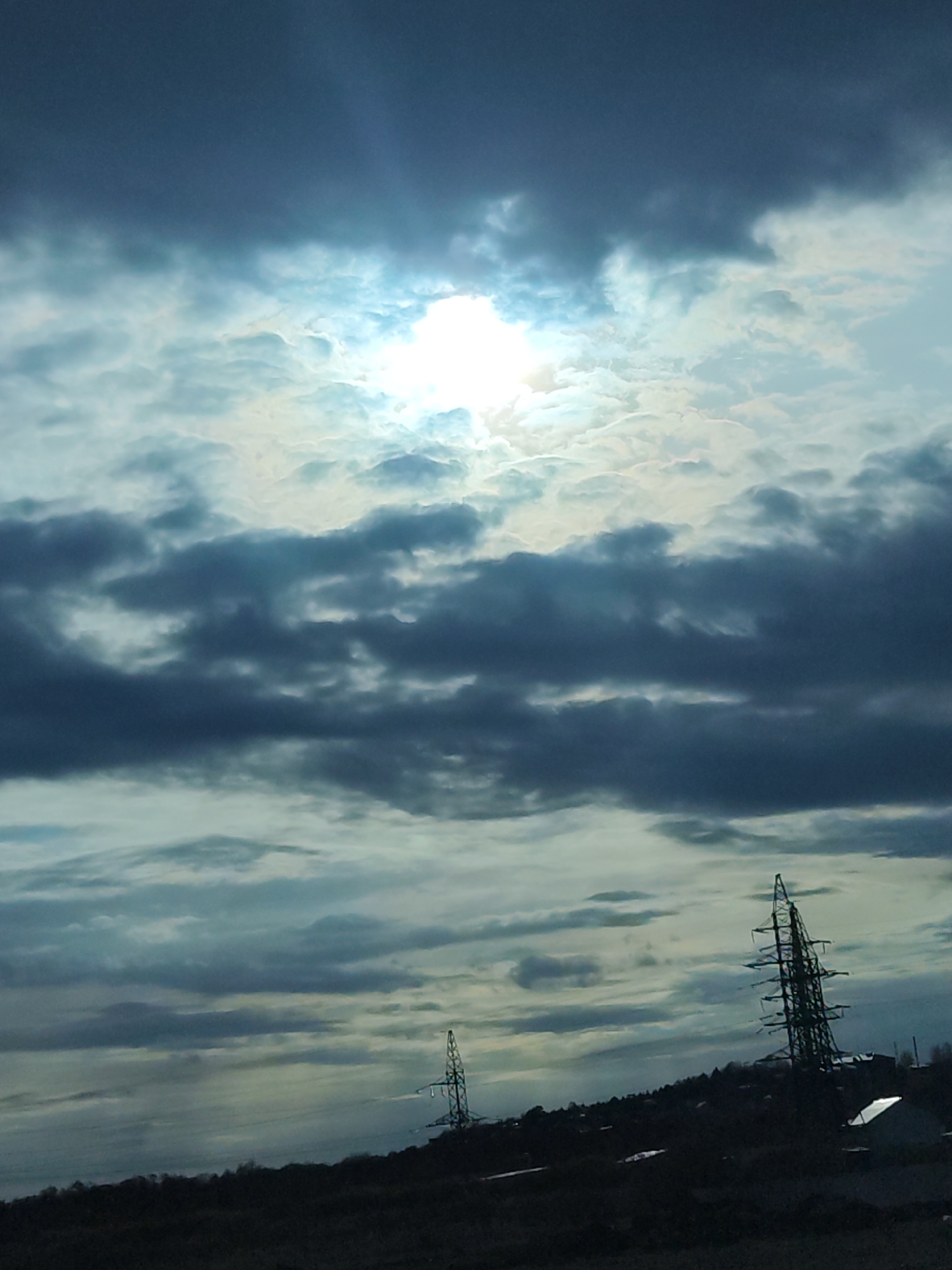 The image depicts a cloudy sky with the sun partially obscured. Below the sky, there is a dark, hilly landscape featuring two electrical transmission towers. In the lower right portion of the image, there are buildings and roofs visible. The photo was taken in Izhevsk, Russia.