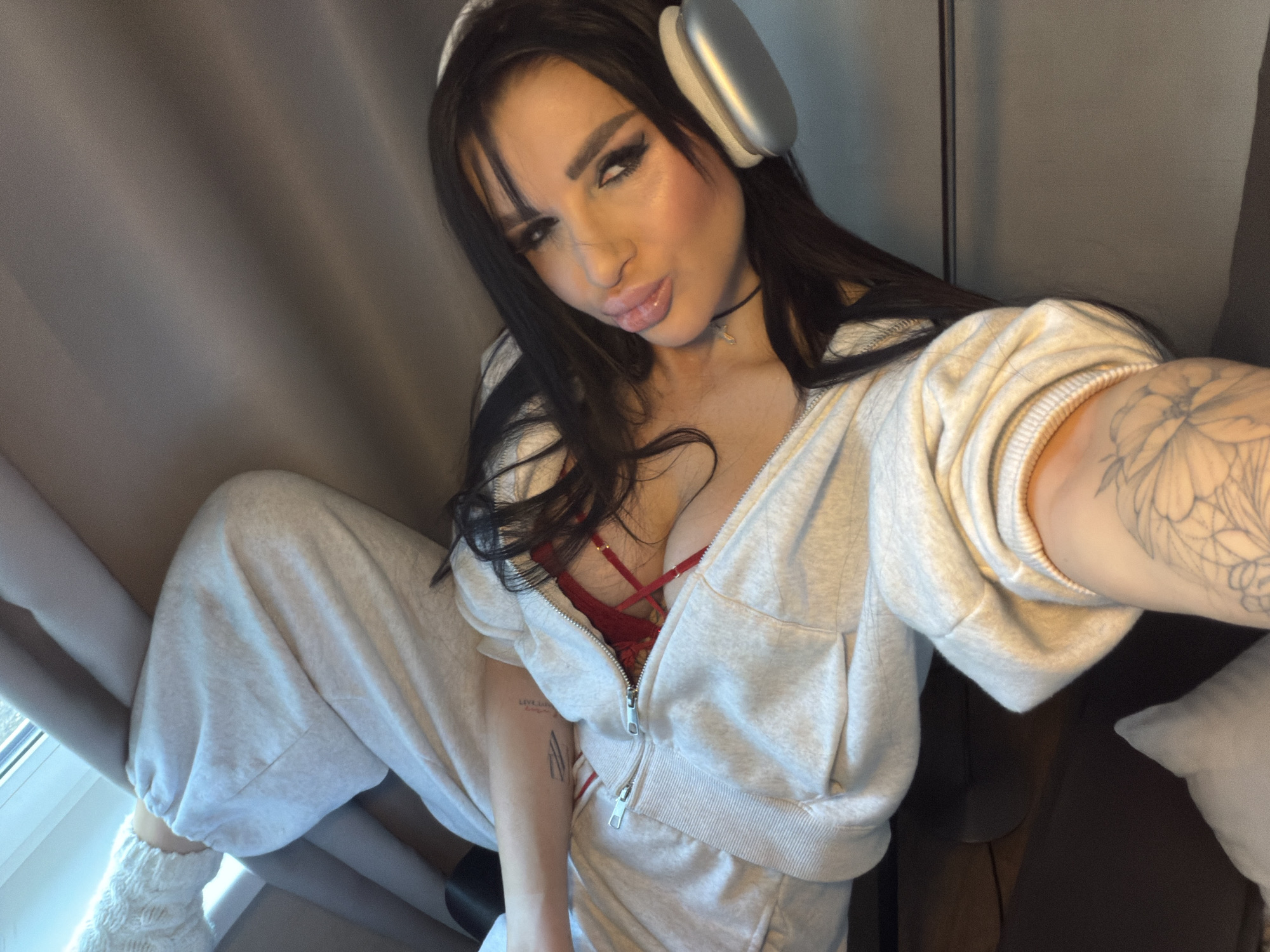 A woman is sitting indoors, wearing a light gray zip-up hooded sweatshirt over a red lace bra and matching sweatpants, and white knitted leg warmers on her left leg. She has headphones around her neck, long dark hair, and is looking at the camera with a slight pout. Her right arm, which has a large floral tattoo, is extended towards the camera as if taking a selfie. Part of a window is visible to the left with a gray curtain partially obscuring the light coming in. The background includes a gray curtain and a dark brown chair or couch, with part of a light gray pillow visible. The location is not specified in the city of Svkh Voronezhskiy, Russia.