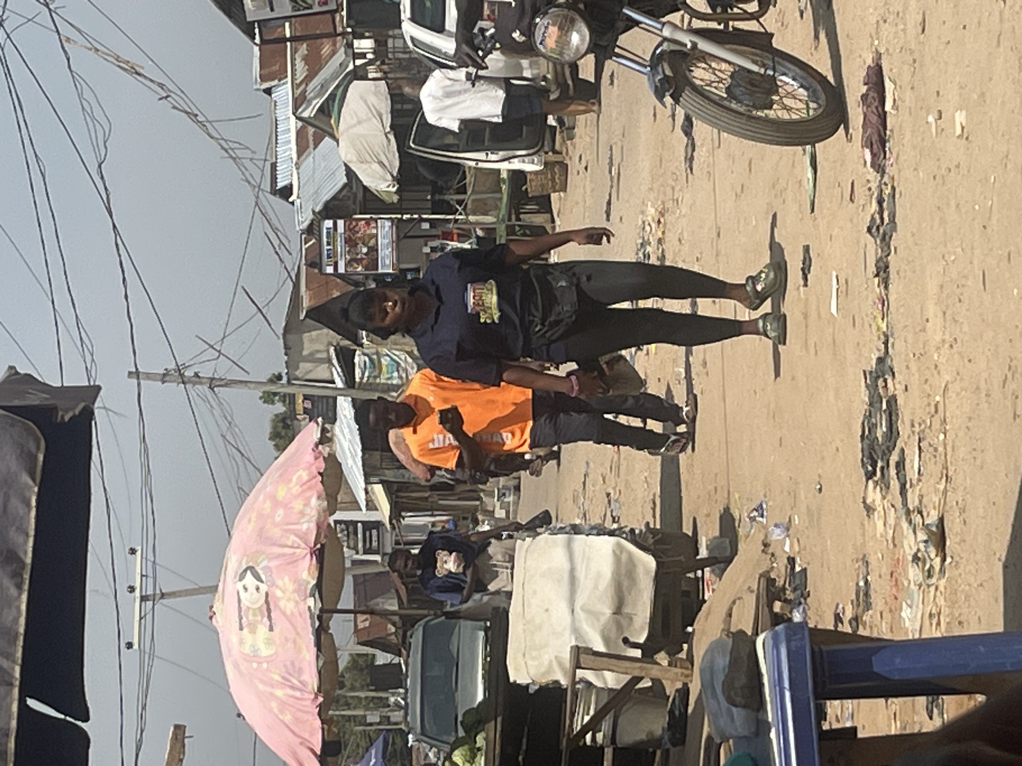 The image depicts a street scene in Abuja, Nigeria, characterized by a bustling marketplace. The foreground is dominated by a dirt path strewn with litter, including plastic bags and discarded clothing. A blue plastic table is partially visible on the left. In the middle ground, several people are walking through the market. A woman in a dark blue t-shirt and leggings, with a fanny pack around her waist, is the most prominent figure, walking towards the viewer. To her left, a man in an orange t-shirt and dark pants is walking away, holding a mobile phone. Behind them, other individuals and market stalls are visible, with corrugated iron roofs and makeshift structures. A pink umbrella with a floral pattern and a cartoonish face is open above some stalls. A white SUV and a motorcycle are also present in the scene. The background shows a line of buildings and utility poles with a complex network of overhead wires, indicating an urban environment. The overall atmosphere is that of a busy, informal market on a sunny day.