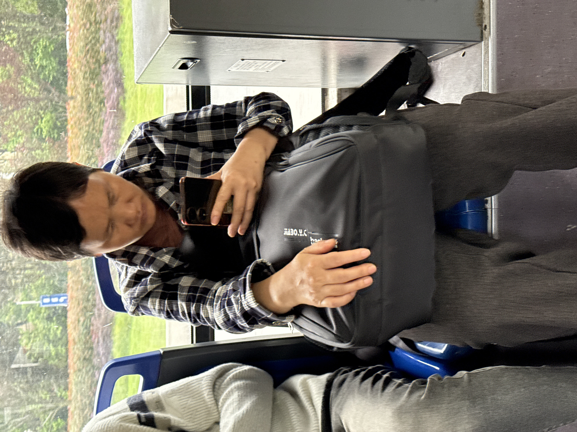 A man is seated on a bus, using a smartphone. He is wearing a plaid shirt, gray pants, and a black backpack. The bus window shows greenery outside, suggesting a moving vehicle. The image orientation is rotated. There's no indication this photo was taken in Jiangjin, China.