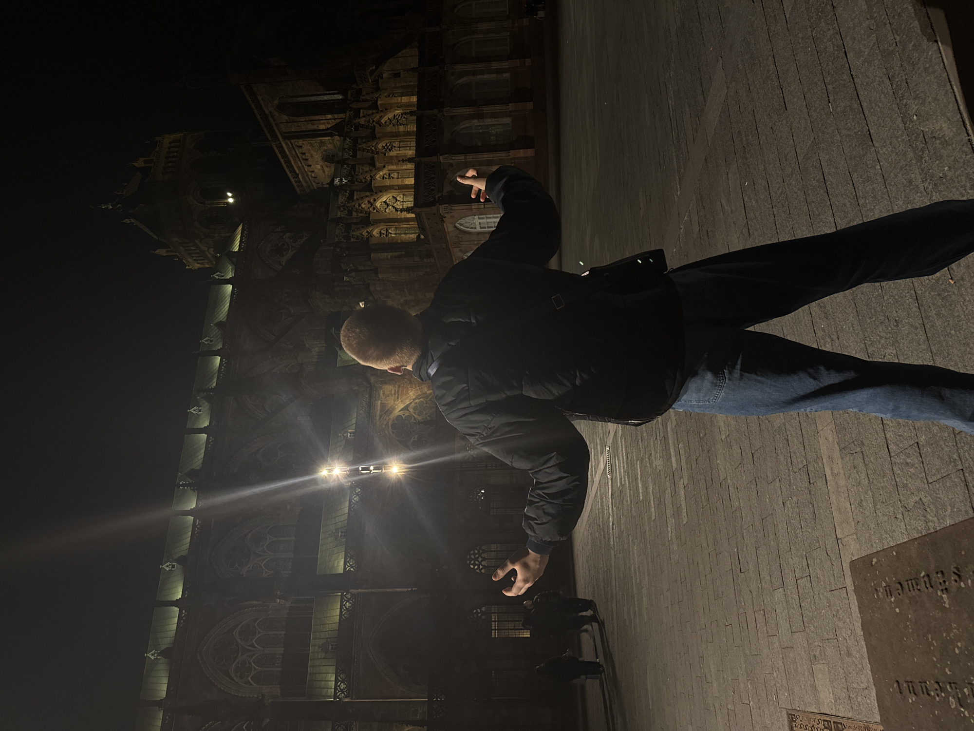 The image captures a person standing on a paved surface at night, positioned in front of Strasbourg Cathedral. The person is wearing a dark jacket and jeans and has their arms raised, with their fingers pointing upwards towards the cathedral. The cathedral is illuminated with lights, highlighting its architectural details. The surrounding environment is dark, with the cathedral dominating the background. A stone marker can be seen in the bottom right corner of the image.