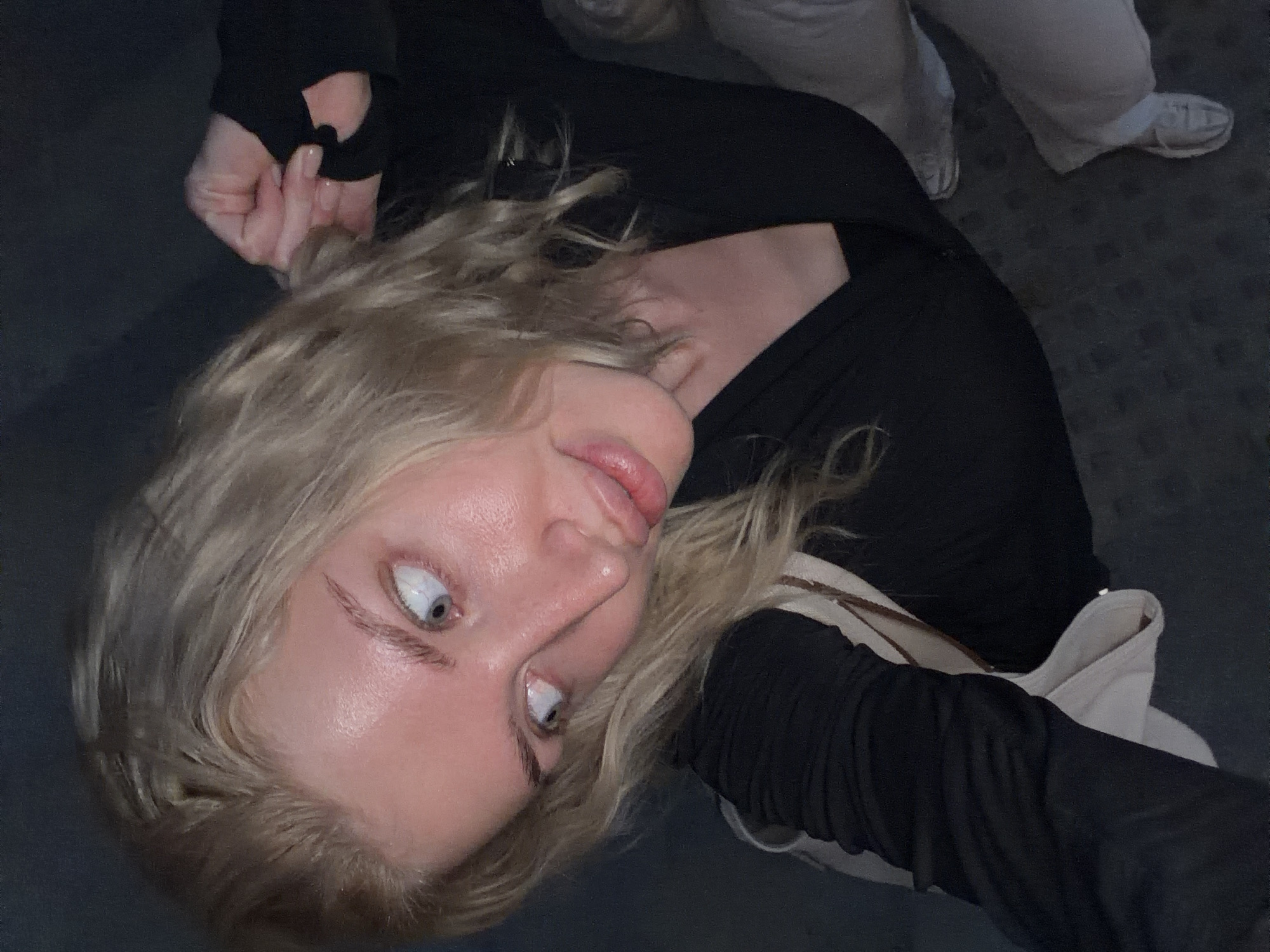 A young woman with blonde, wavy hair and fair skin takes a selfie. She is wearing a black long-sleeved shirt and a light-colored shoulder bag. Her eyes are open wide, and her lips are slightly parted. Her fingers are clutching her hair behind her head. In the background, a person is wearing light-colored pants and white sneakers is visible. The ground is dark gray. The location is not identifiable as being in Belgrade, Serbia, from the image alone.