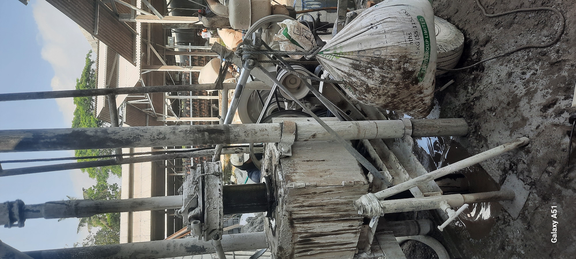 The image depicts an outdoor scene in Ploso, Indonesia, likely a construction or industrial site. In the foreground, a drilling rig is partially visible, heavily coated in mud and grime. A large, white sack, also stained with mud, is suspended by a rope, displaying text that includes "55.115LBS." and "GROUP LIMITED." Several thick, weathered metal pipes or supports rise vertically, forming part of the rig's structure. Muddy water pools on the ground around the base of the machinery. In the background, a shed-like structure with a corrugated metal roof is visible, and further behind that, rows of what appear to be clay bricks or building materials are stacked. A person wearing a white cap and dark clothing is partially visible, appearing to be operating or attending to the drilling equipment. Another person in an orange shirt is seen in the distance within the shed structure. The overall environment suggests an active work area, with evidence of manual labor and ongoing construction or material processing. The sky is partly cloudy. The bottom left corner displays "Galaxy A51."