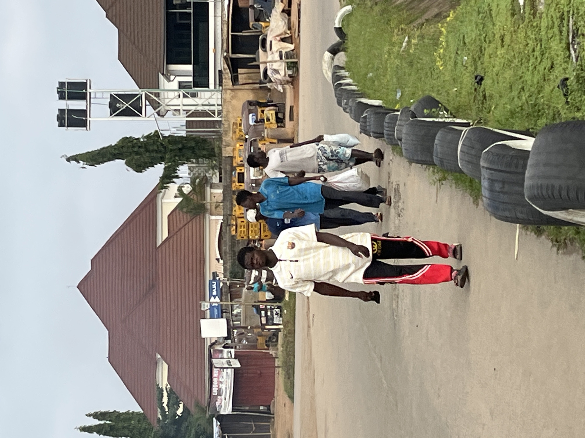 In Abuja, Nigeria, a group of four young men walk along a paved sidewalk adjacent to a road. The man in the foreground wears a white, short-sleeved polo shirt with horizontal stripes and red and black track pants. He is holding a dark object in his right hand. Behind him, three other men walk in the same direction. The second man from the front wears a blue t-shirt and black pants, carrying a white plastic bag in his right hand. The third man wears a grey t-shirt and patterned shorts, also carrying a white plastic bag. The fourth man, partially visible, wears a white cap and a light-colored shirt.
The environment features a street with a line of old tires acting as a barrier along the sidewalk, bordering a patch of green grass. In the background, buildings with red-tiled roofs are visible, along with trees and utility structures, including a tall metal tower supporting water tanks. Several vehicles, including what appear to be three-wheeled auto-rickshaws, are parked or driving on the road in the middle ground. A sign with the word "BAJAJ" is visible on one of the structures in the background. The sky is overcast, suggesting a cloudy day.
