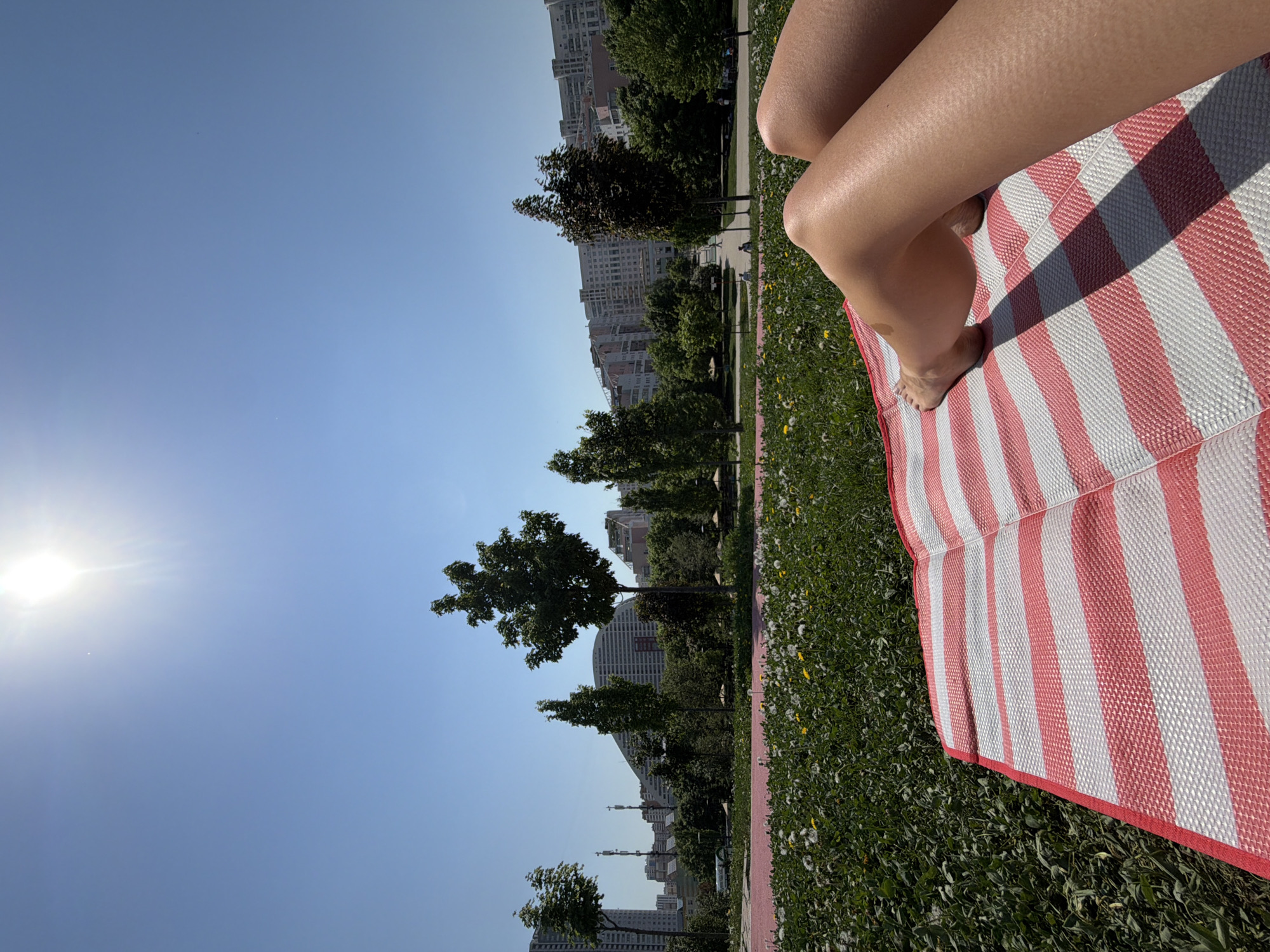 The image shows a person lying on a red and white striped mat in a grassy area on a sunny day. The person's legs are visible on the mat. The background features trees and buildings, suggesting an urban park setting. Based on the architectural style of the buildings in the background, the location is likely Moscow, Russia.
