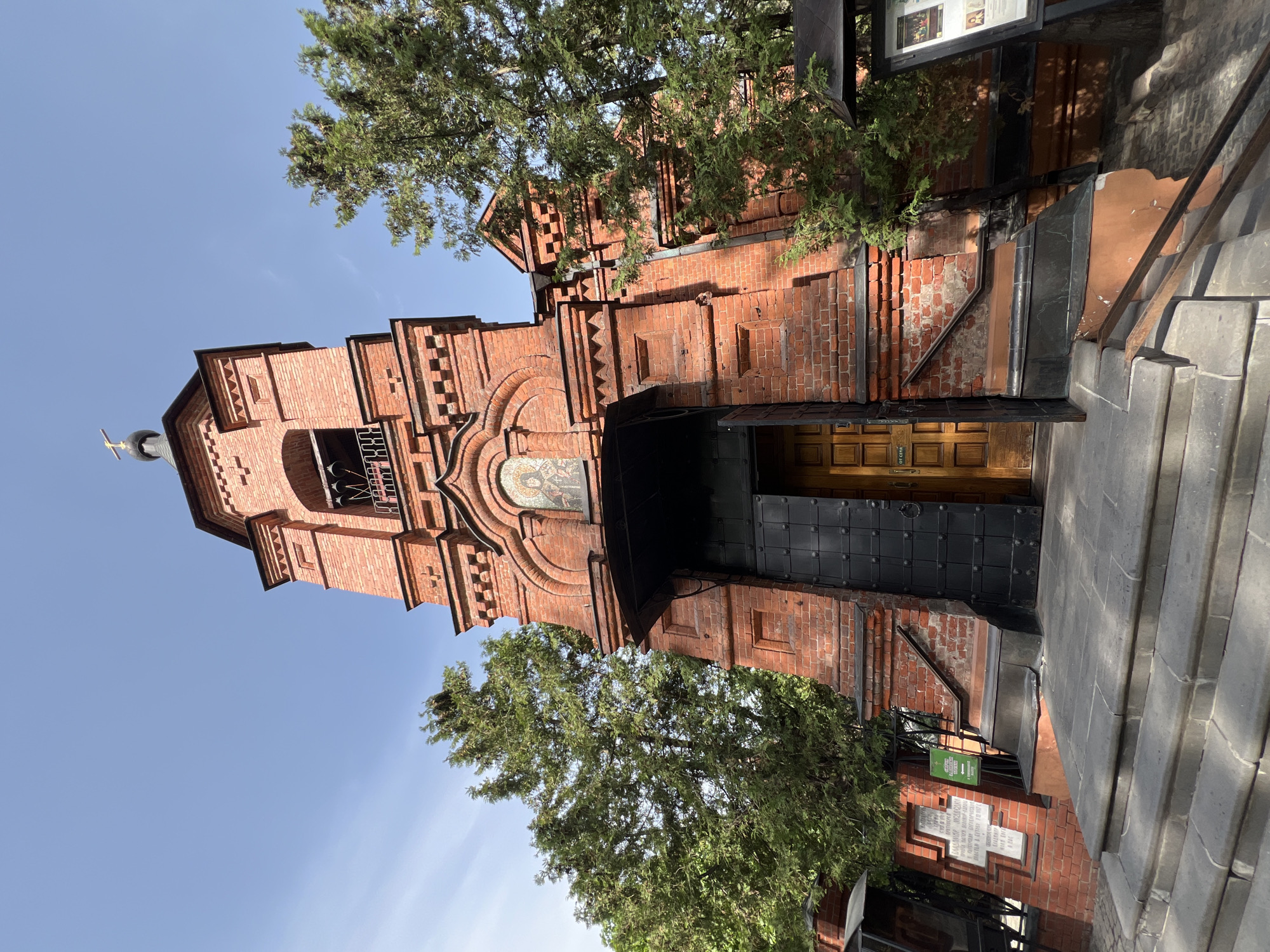 A low-angle shot shows the red brick facade of the Church of the Assumption of the Blessed Virgin Mary in Khimki, Russia. The church features a bell tower topped with a cross. A mosaic decorates the entrance arch. The church door is partially open, revealing a glimpse of the interior. Stone steps lead up to the entrance. Green trees flank the structure, obscuring parts of the view. A sign and plaques are visible near the base of the building. The sky is light blue.