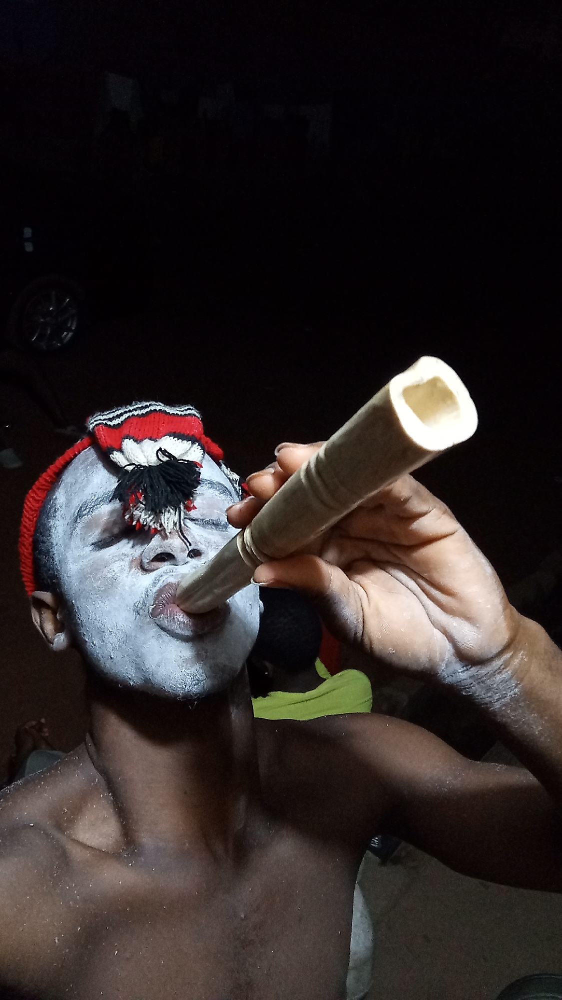 A man with white face paint is blowing into a light-colored, cylindrical object, possibly a horn or flute. He wears a red and white headband with black tassels. The photo appears to be taken at night or in a dimly lit place. A person wearing a yellow shirt is partially visible in the background. A car tire is visible in the background on the left. The location is Onitsha, Nigeria.
