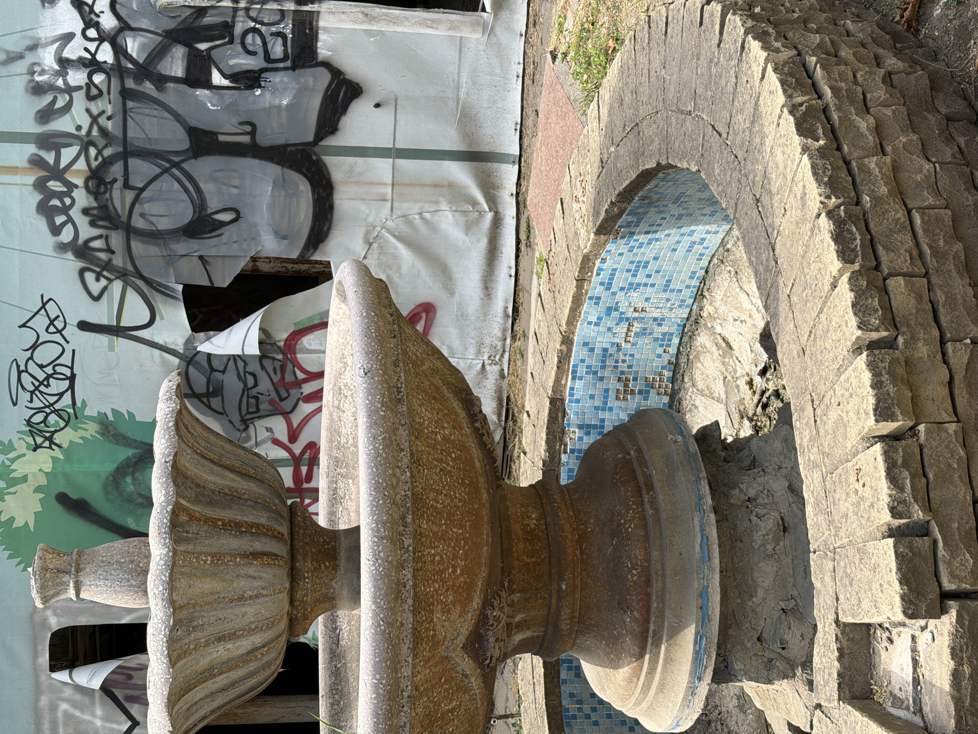 The image shows a two-tiered, weathered stone fountain situated in an outdoor setting. The fountain is the central subject, with its upper basin featuring decorative fluting and a finial on top. The lower basin is wider and also exhibits signs of age and wear. The fountain is surrounded by a circular stone wall built with rough-hewn blocks. Inside the wall, a tiled basin with small blue and white tiles is visible, though the fountain itself appears dry and neglected. Behind the fountain, a wall covered in graffiti is prominent. The graffiti is abstract and colorful, with various tags and lettering. Torn white sheeting is attached to the graffiti wall in some areas, revealing glimpses of what might be construction or a dilapidated structure underneath. The ground around the fountain is covered in dry leaves and patchy grass. The overall impression is one of disuse and urban decay. The location is identified as Rostov-on-Don, Russia.